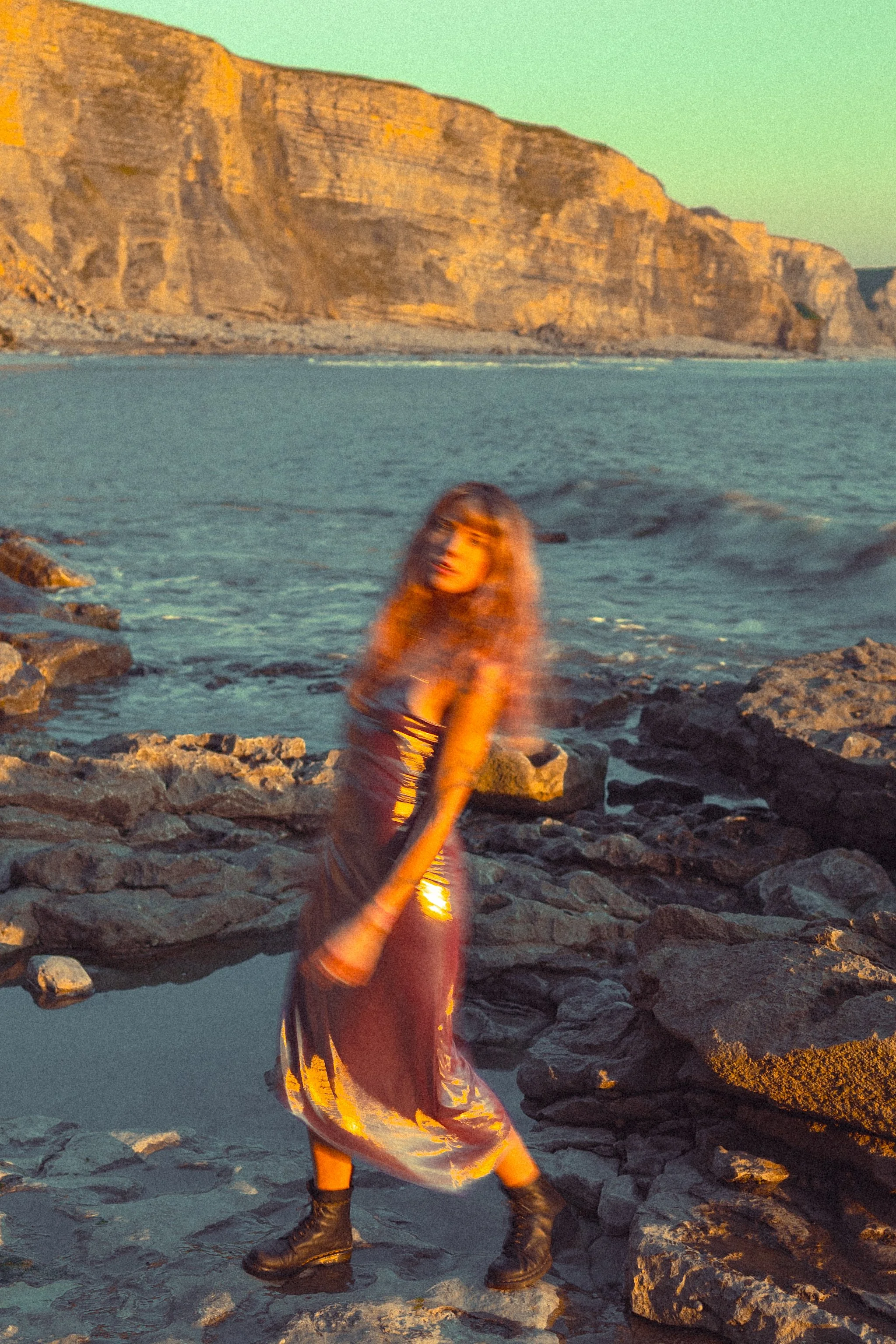 A woman with long, curly hair wearing a dark dress with shiny, reflective material and black combat boots standing on rocky coastline with cliffs in the background during sunset.