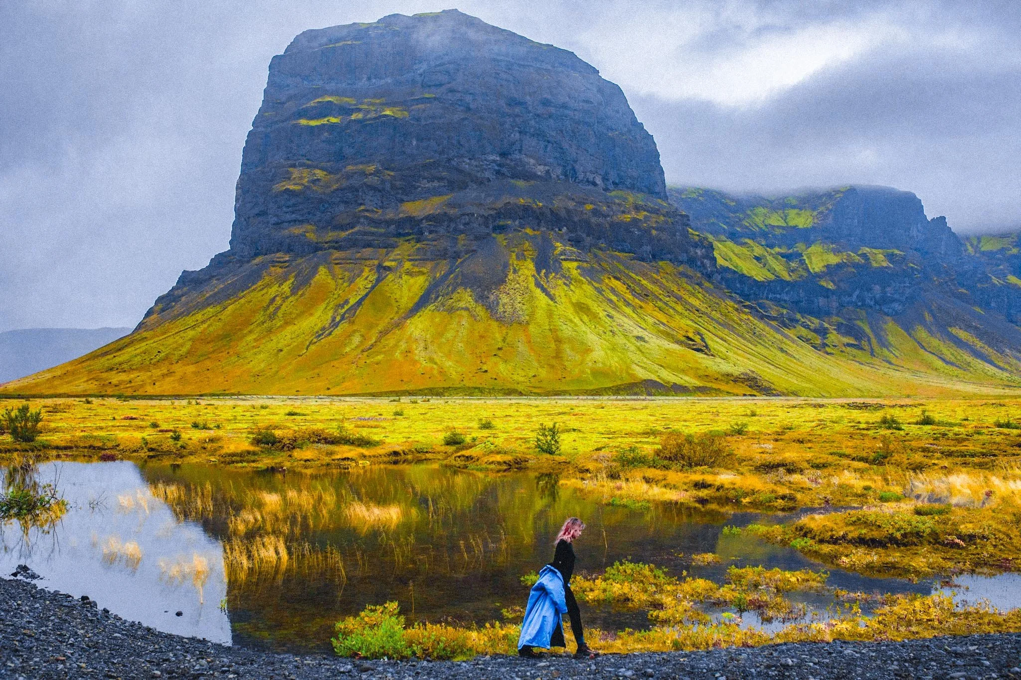 Woman walking beside reflective water with large mountain rising behind her