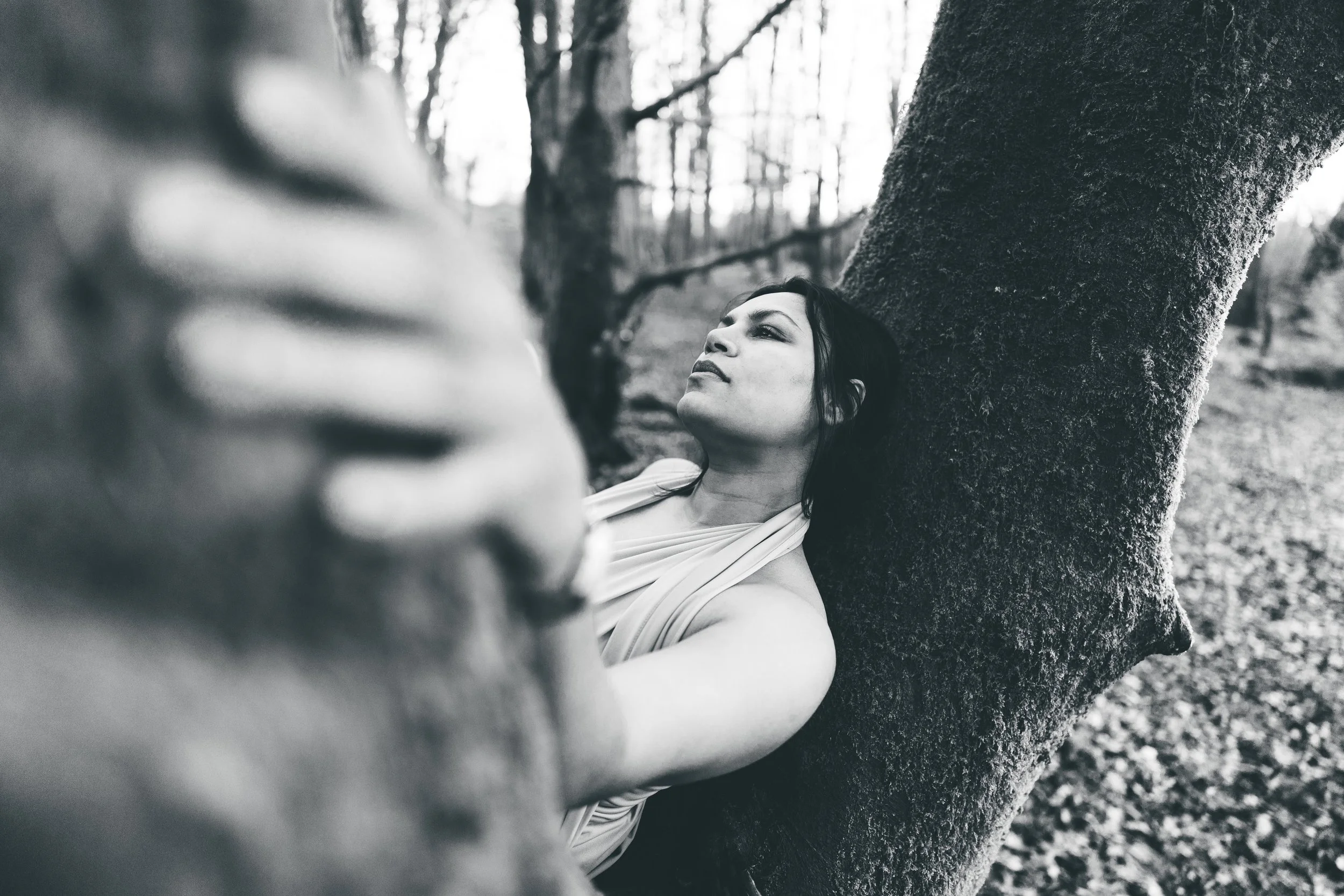 A black-and-white photo of a woman leaning against a tree in a forest, gazing upward with a serene expression.
