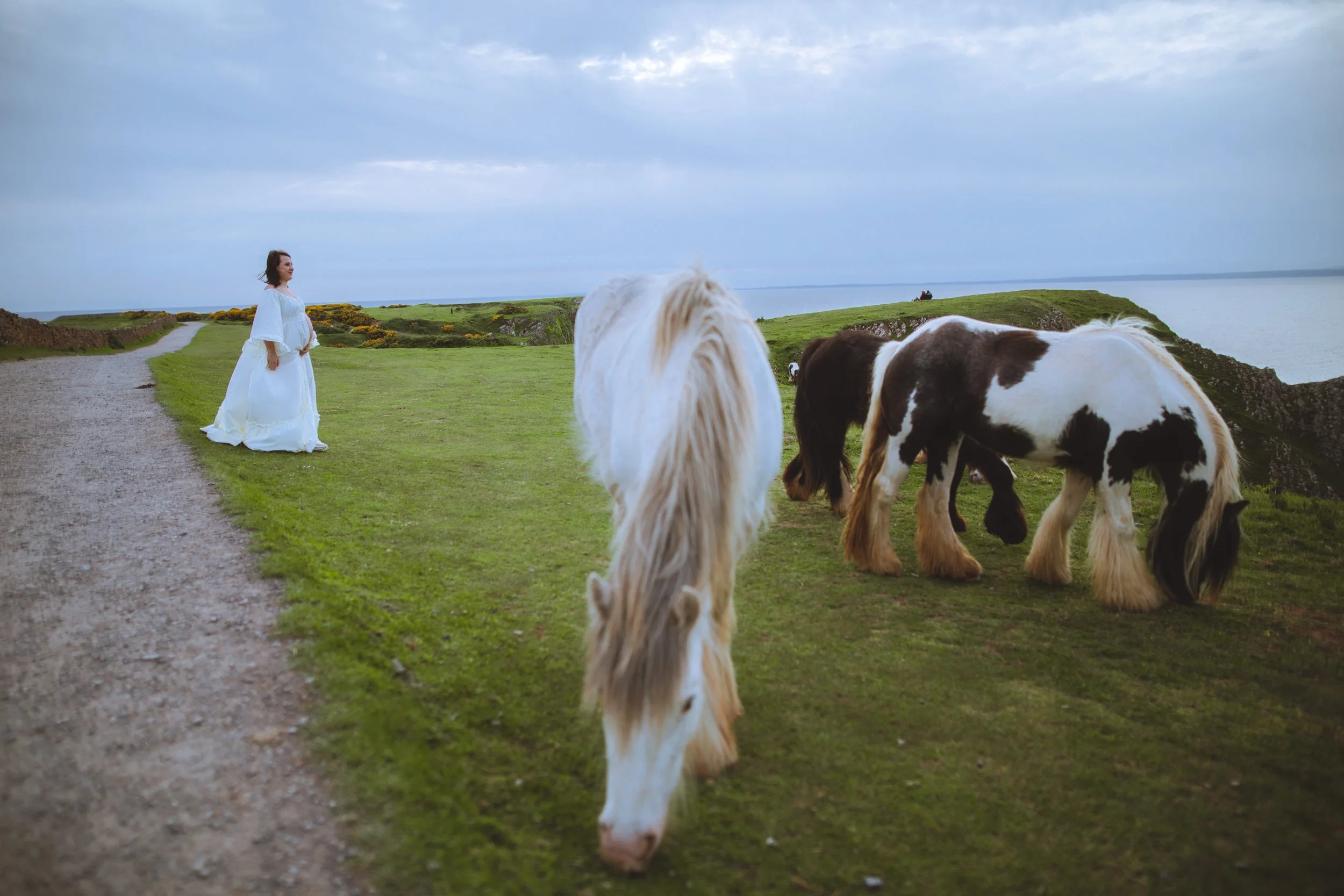 A woman in a long white dress standing on a grassy path near grazing horses with a lake in the background and cloudy sky overhead.