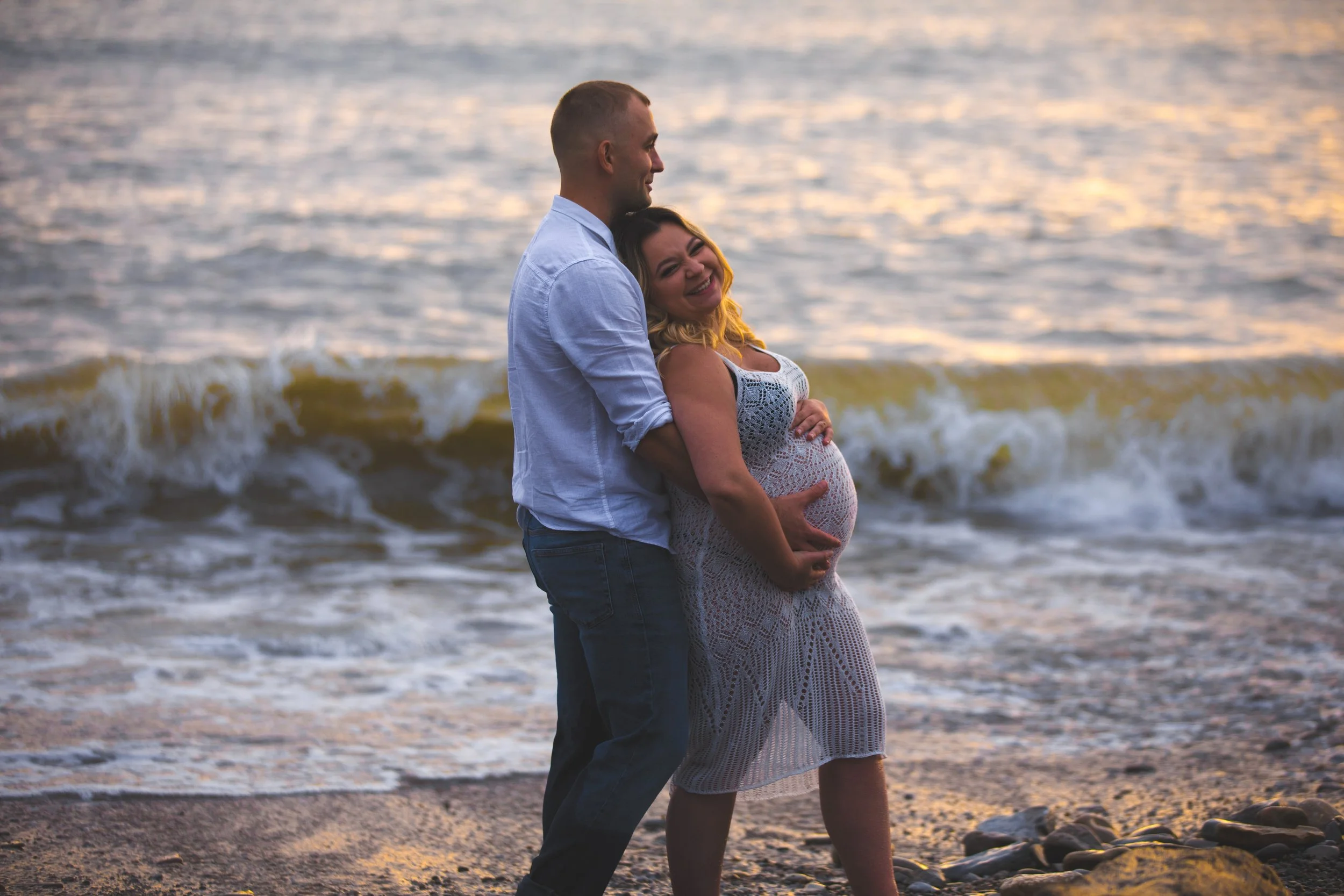 A happy pregnant woman and her partner standing on the beach at sunset, embracing with the ocean in the background.