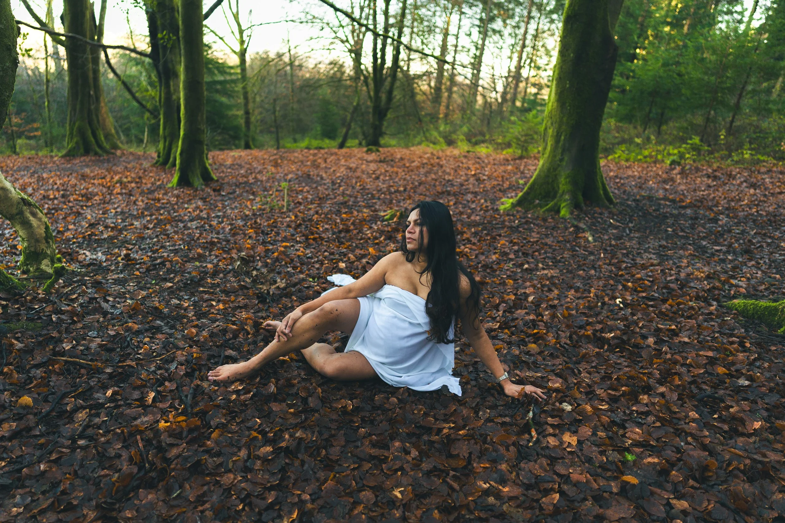 A woman with long dark hair, wearing a white towel, sitting on a bed of fallen leaves in a forest during daytime.