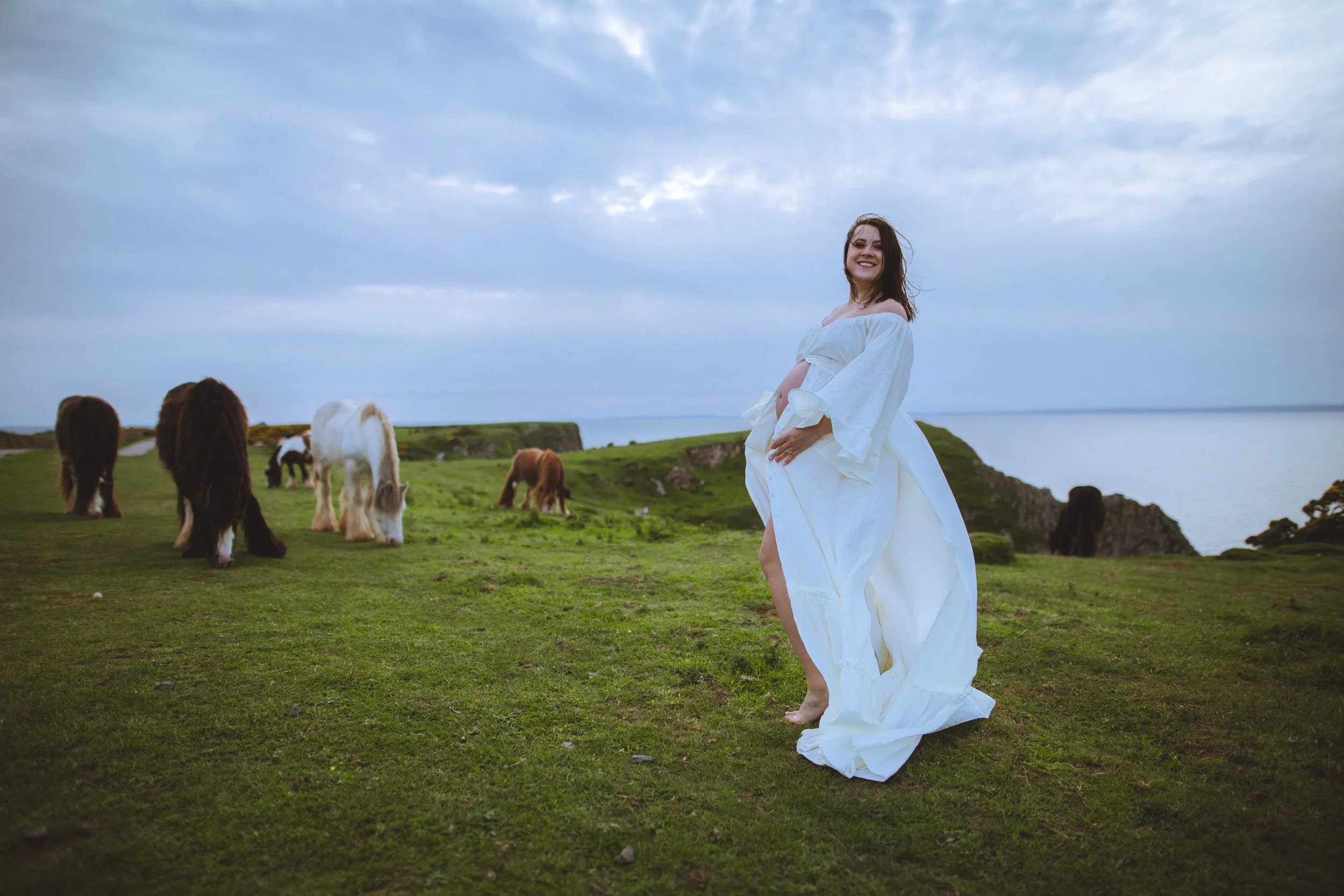 Pregnant woman in a white flowing dress smiling on a grassy cliff with cows grazing in the background, overlooking the ocean.