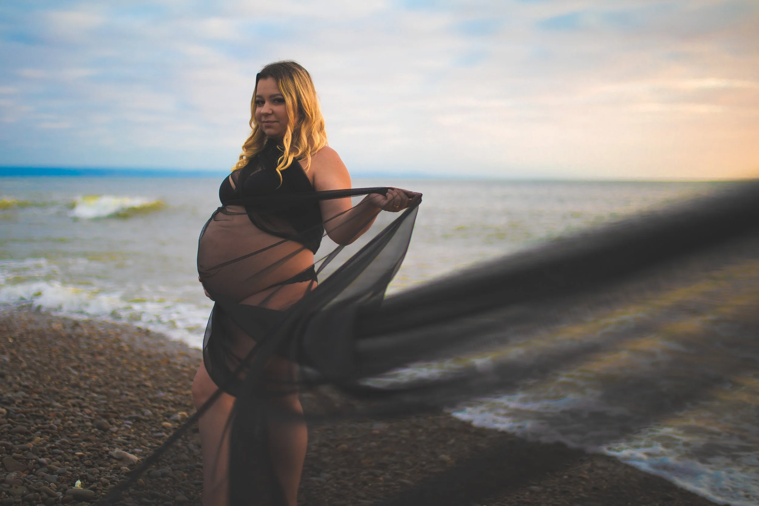 A pregnant woman in black lingerie and a sheer black wrap posing on a pebbled beach during sunset, with the ocean behind her.