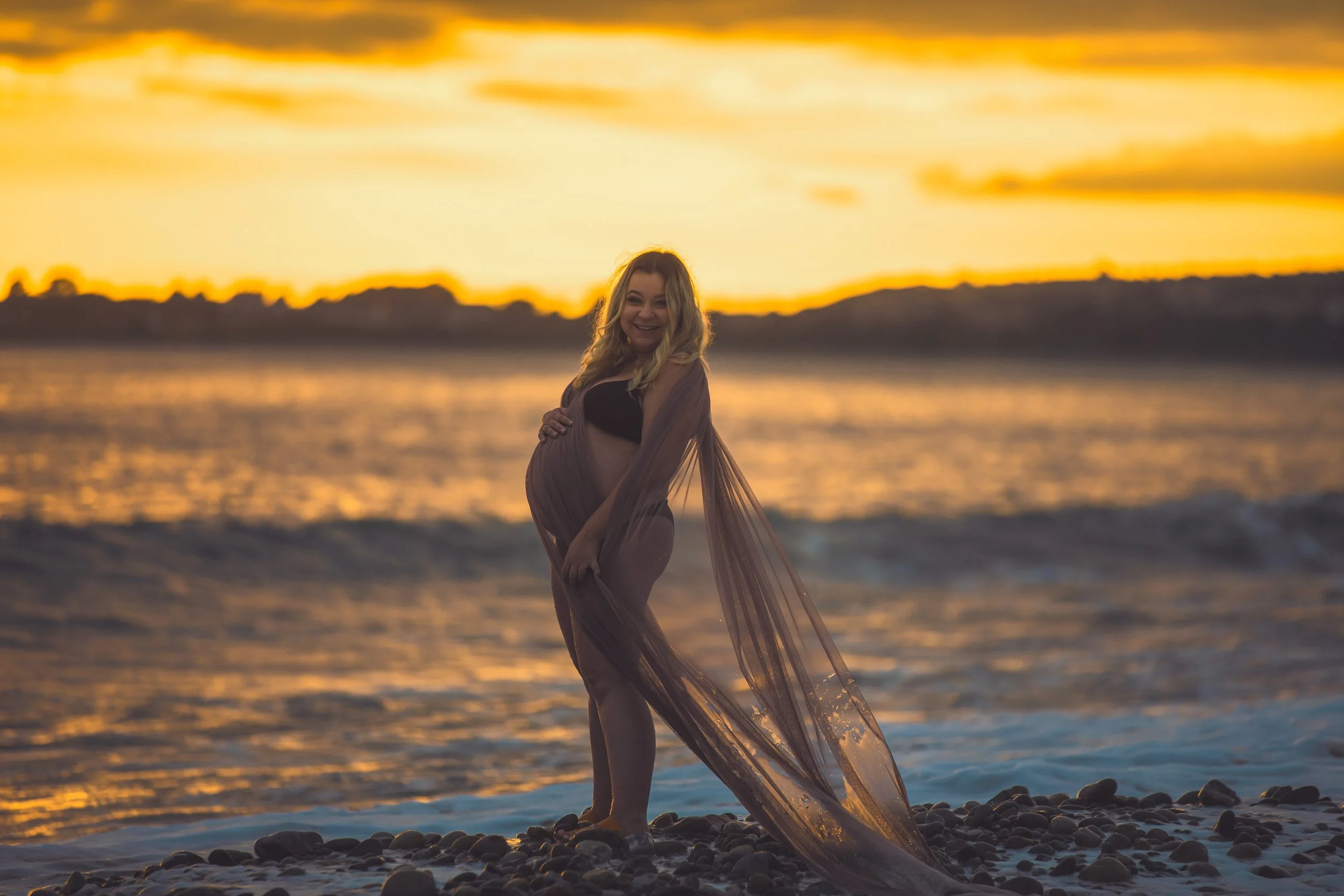 A pregnant woman is standing on a rocky beach at sunset, smiling and holding her belly, with water and hills in the background.