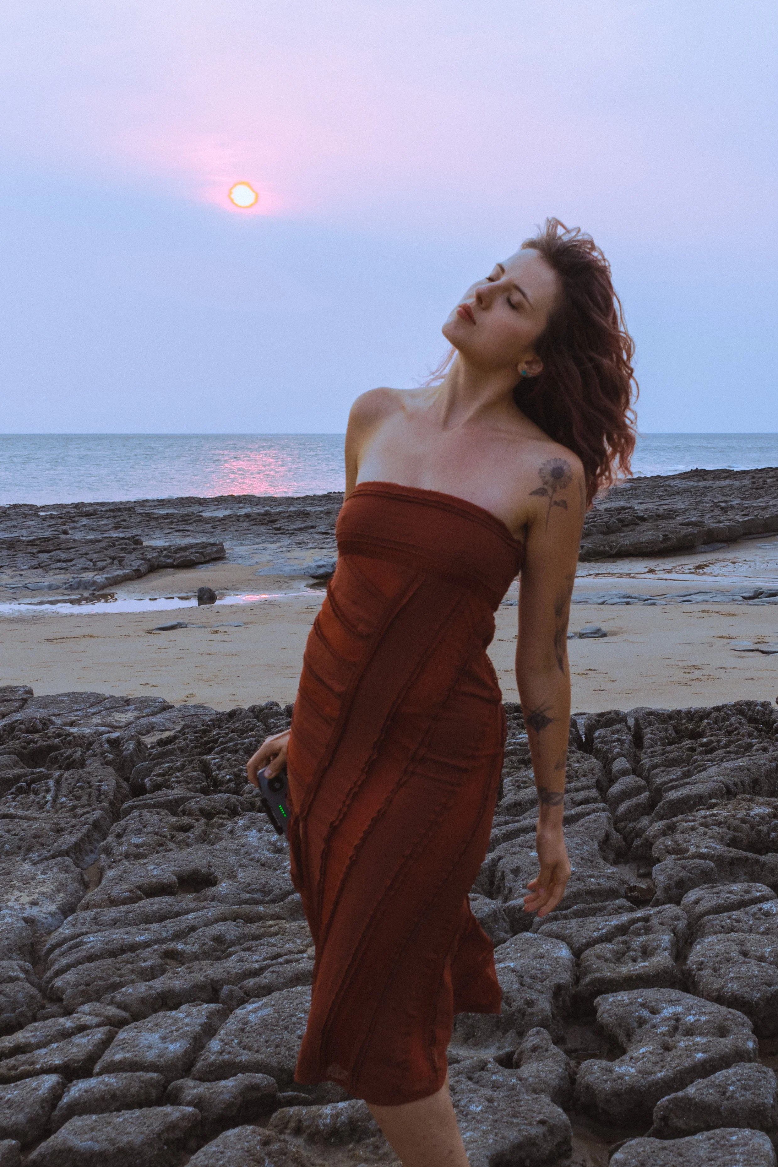 A woman with curly hair wearing a strapless brown dress stands on rocky terrain at the beach during sunset with her eyes closed.