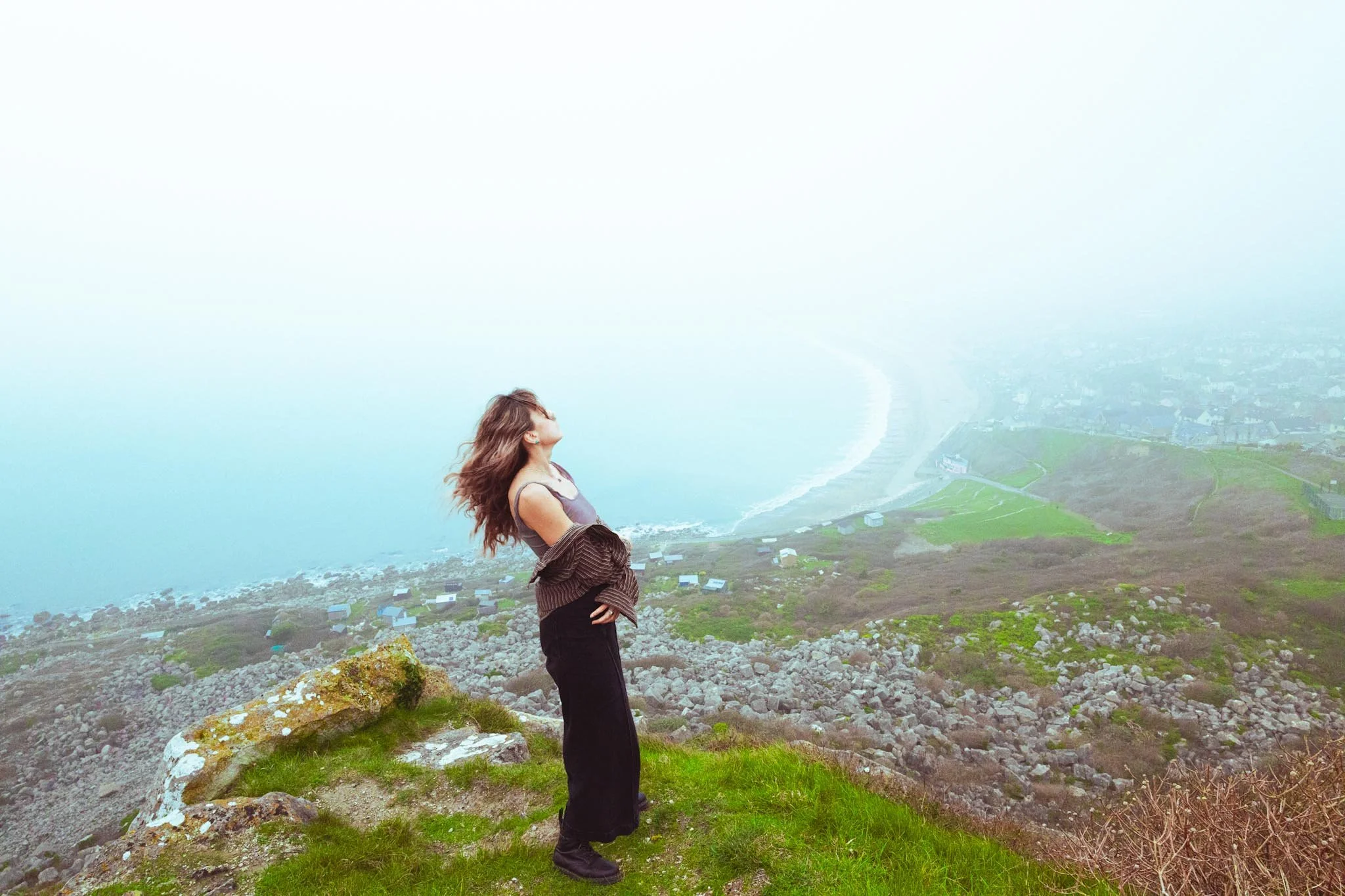Woman standing on grassy cliff overlooking misty coastline and curved shoreline below