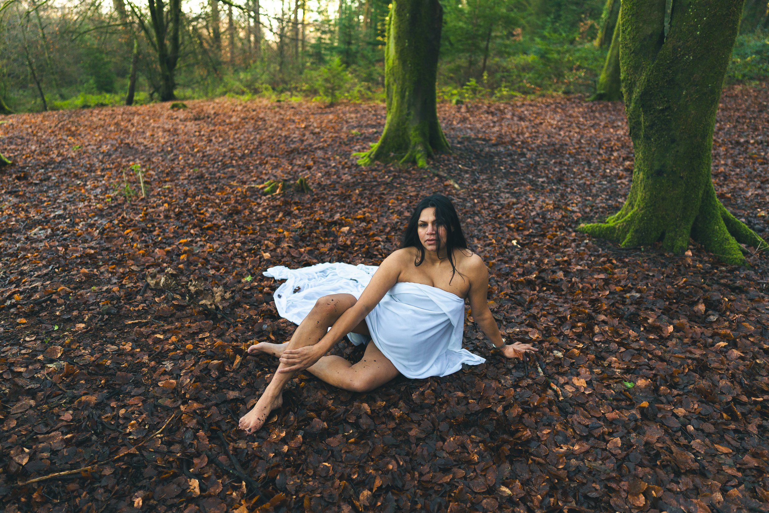 A woman wearing a white dress sitting on a bed of fallen autumn leaves in a forest