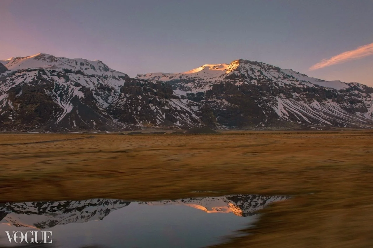 Snow-covered mountains reflected in a calm lake at sunset, with golden grass in the foreground.