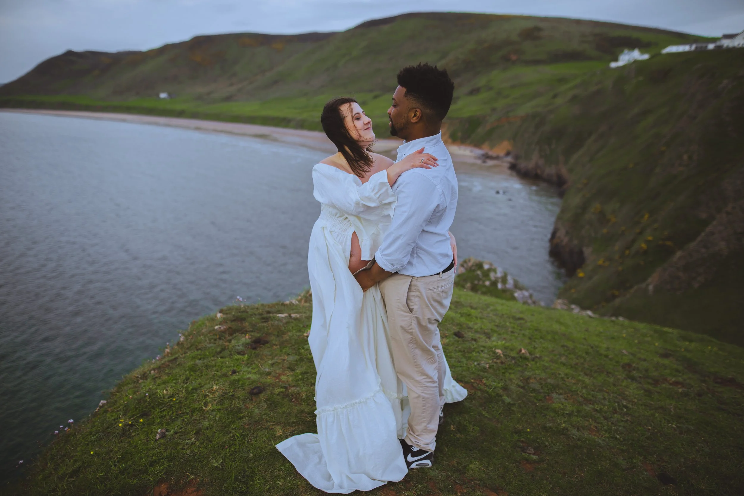 A couple standing on a grassy cliffside by a body of water, with hills in the background. The woman is wearing a white dress, and the man is dressed in a light blue shirt and beige pants. They are embracing and looking at each other affectionately.