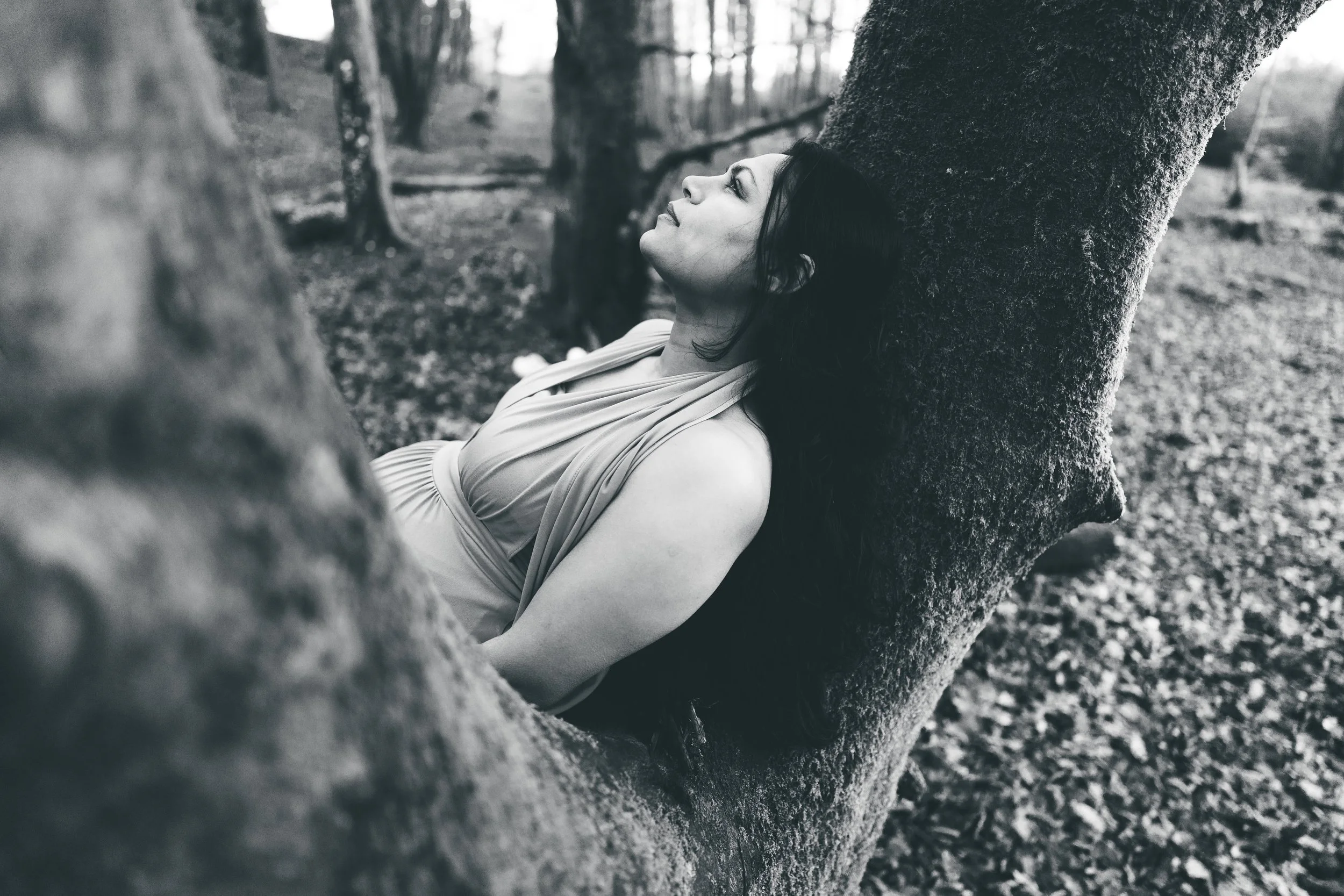 A woman with dark hair reclining against a tree in a wooded area, eyes closed, appearing relaxed and peaceful.