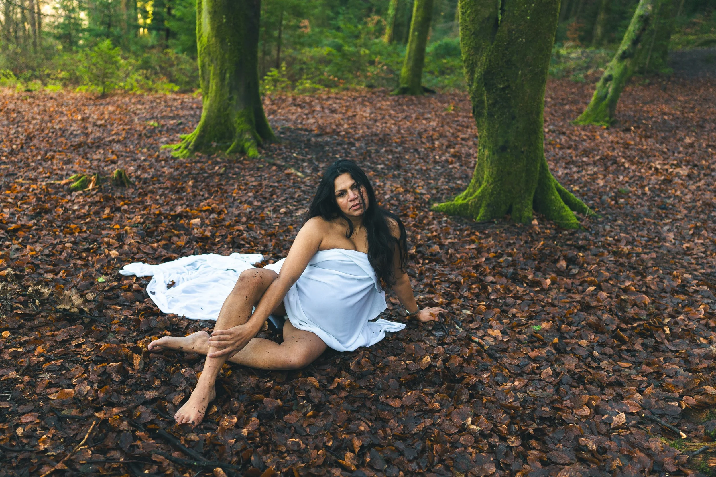 A woman in a white dress sitting on a bed of brown leaves in a forest with trees covered in green moss.