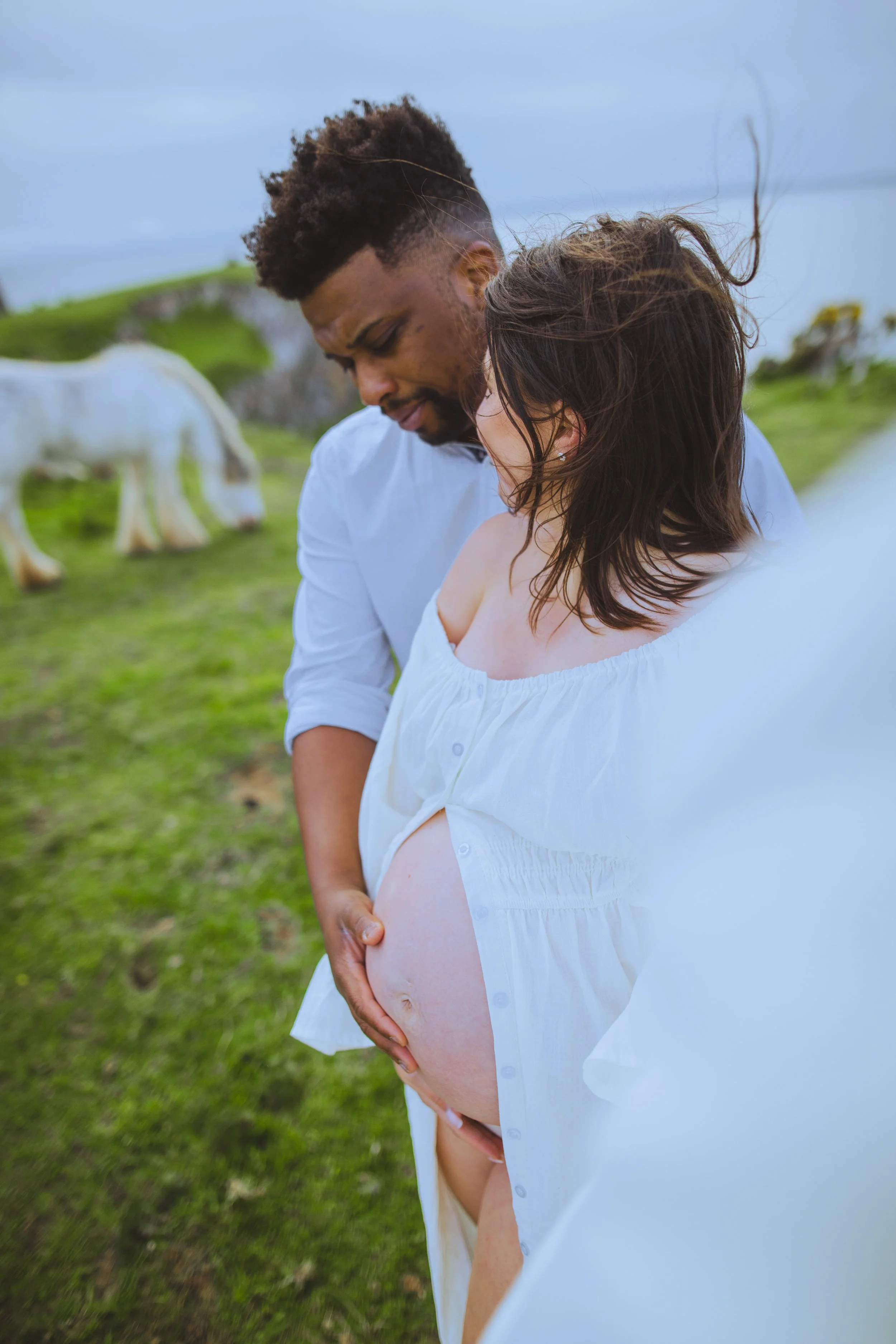 A pregnant woman in a white dress outdoors with a man, touching her belly as they stand on a grassy field with a horse in the background.