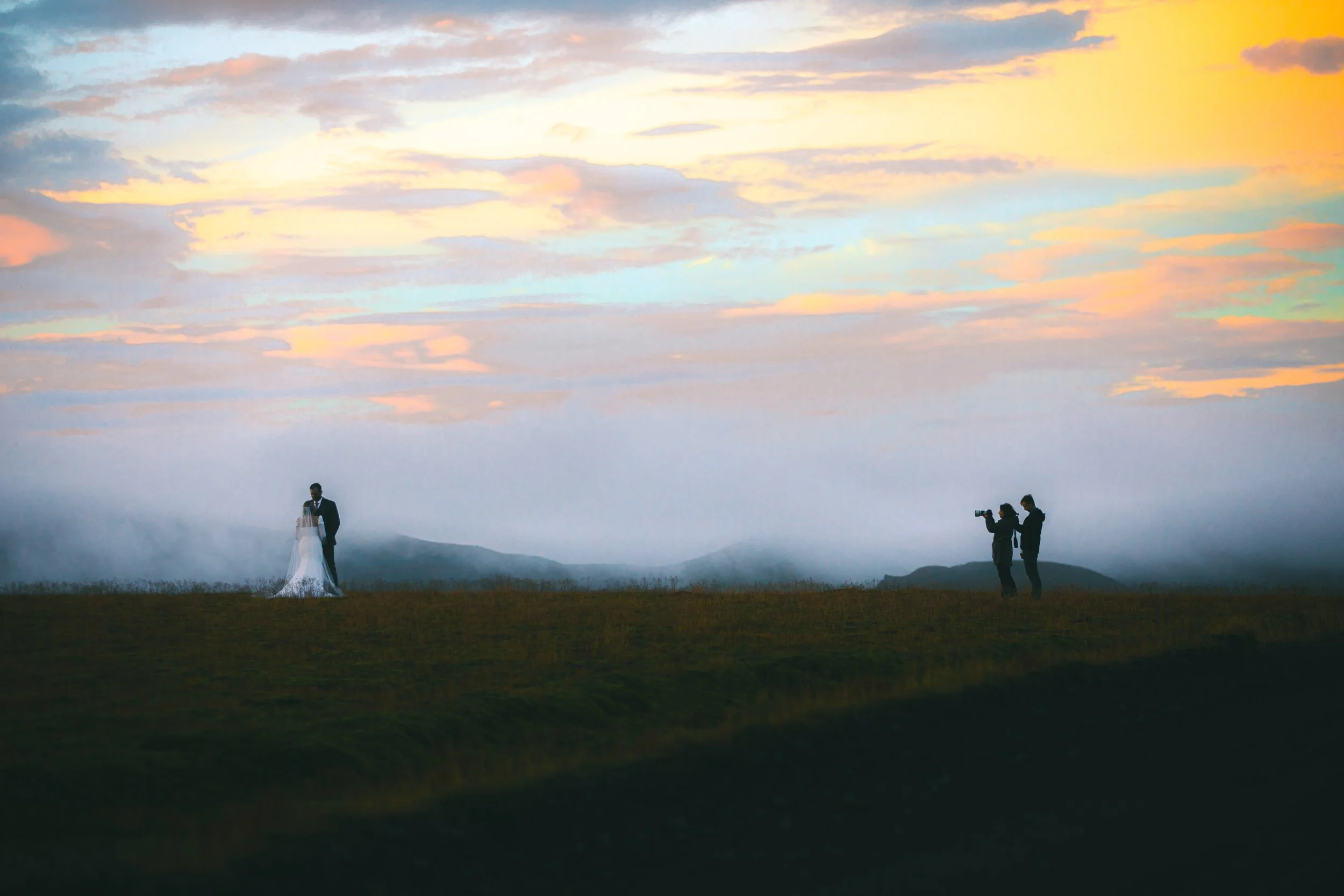 A bride and groom standing outdoors at sunset with two photographers capturing the moment.