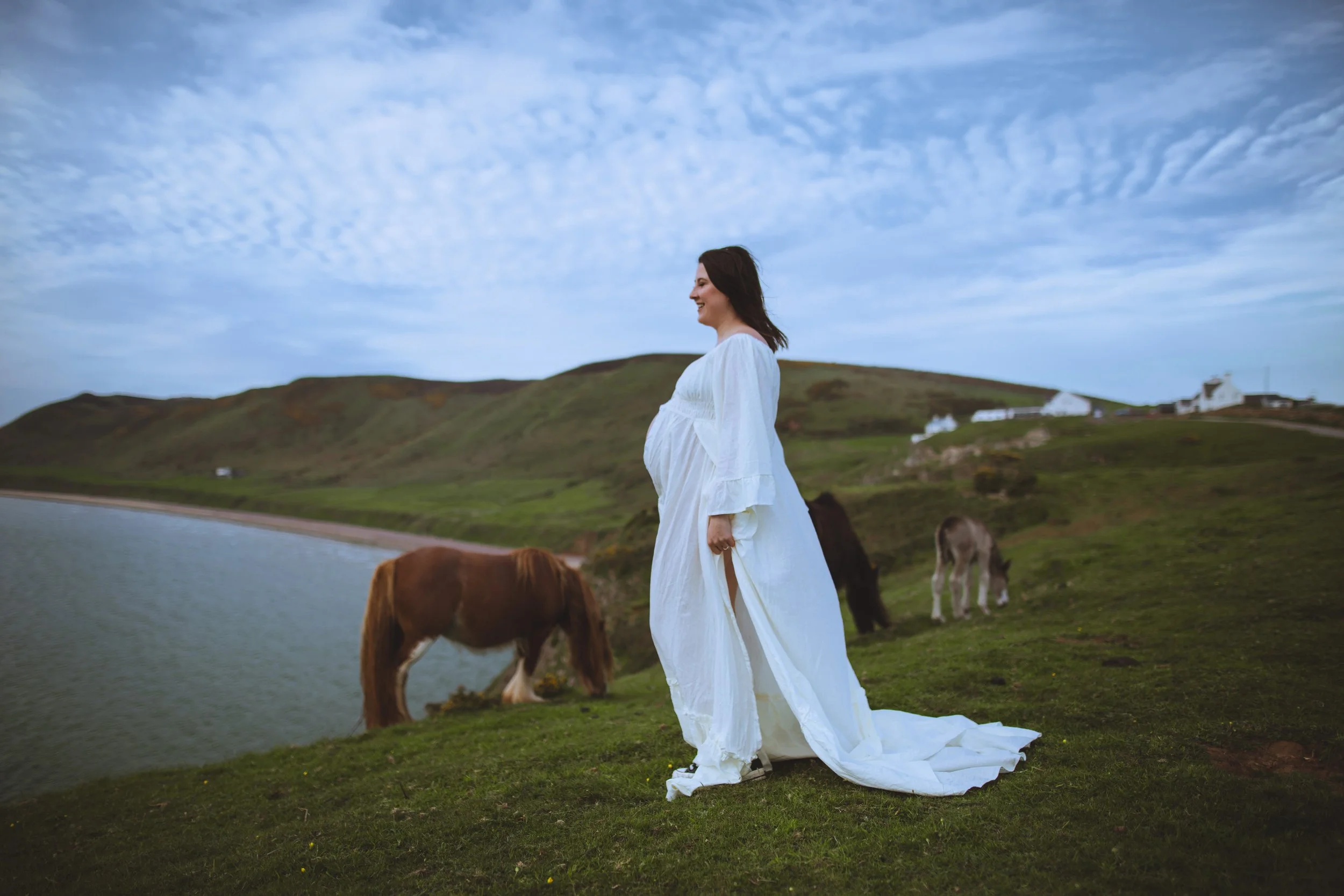 Pregnant woman in a white dress standing on green grass near a pond with grazing horses, hills, and a partly cloudy sky in the background.