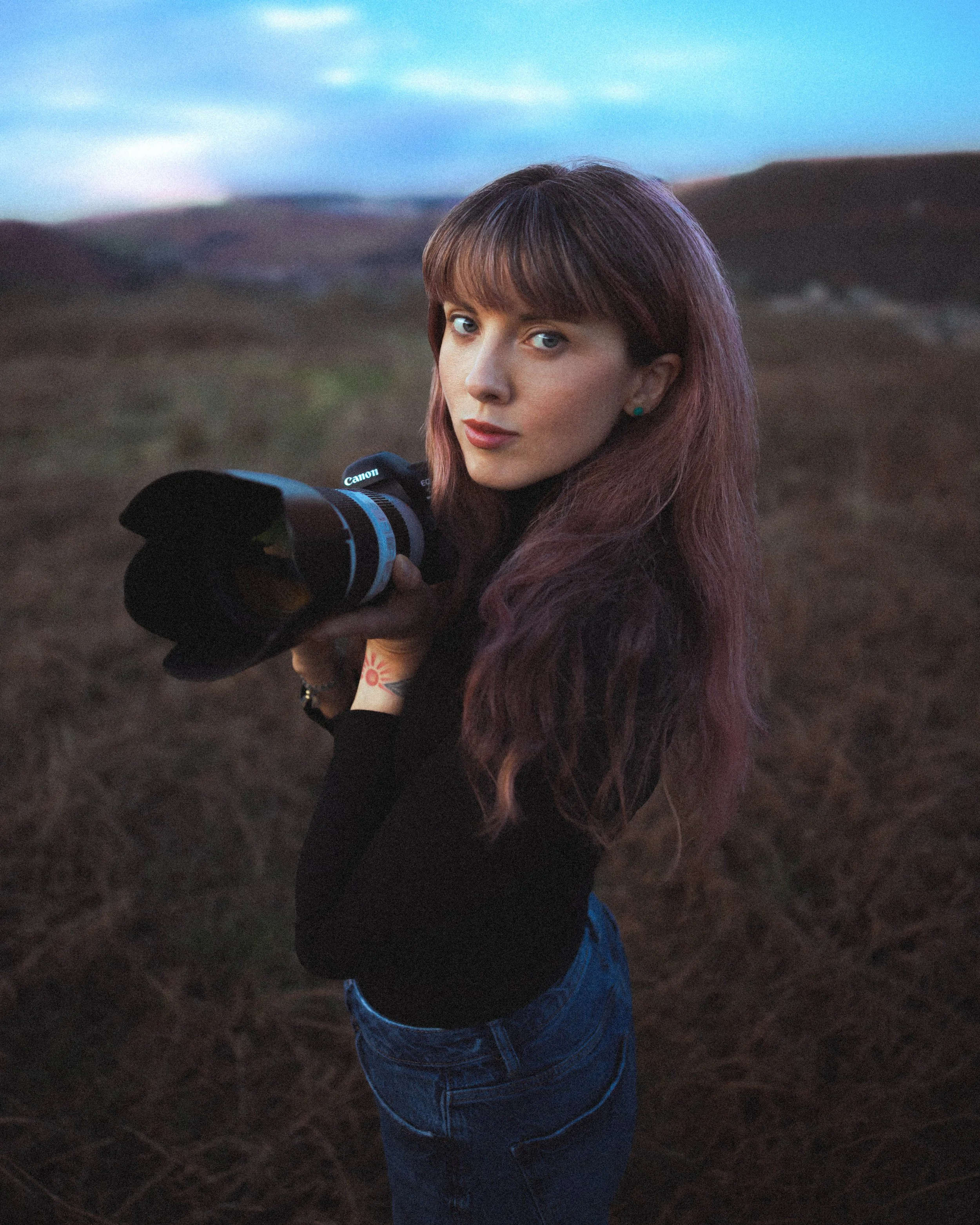 A young woman with long, wavy, reddish-brown hair holding a camera with a large lens outdoors during twilight. She is wearing a black top and blue jeans, standing in a landscape with rolling hills and a partly cloudy sky in the background.