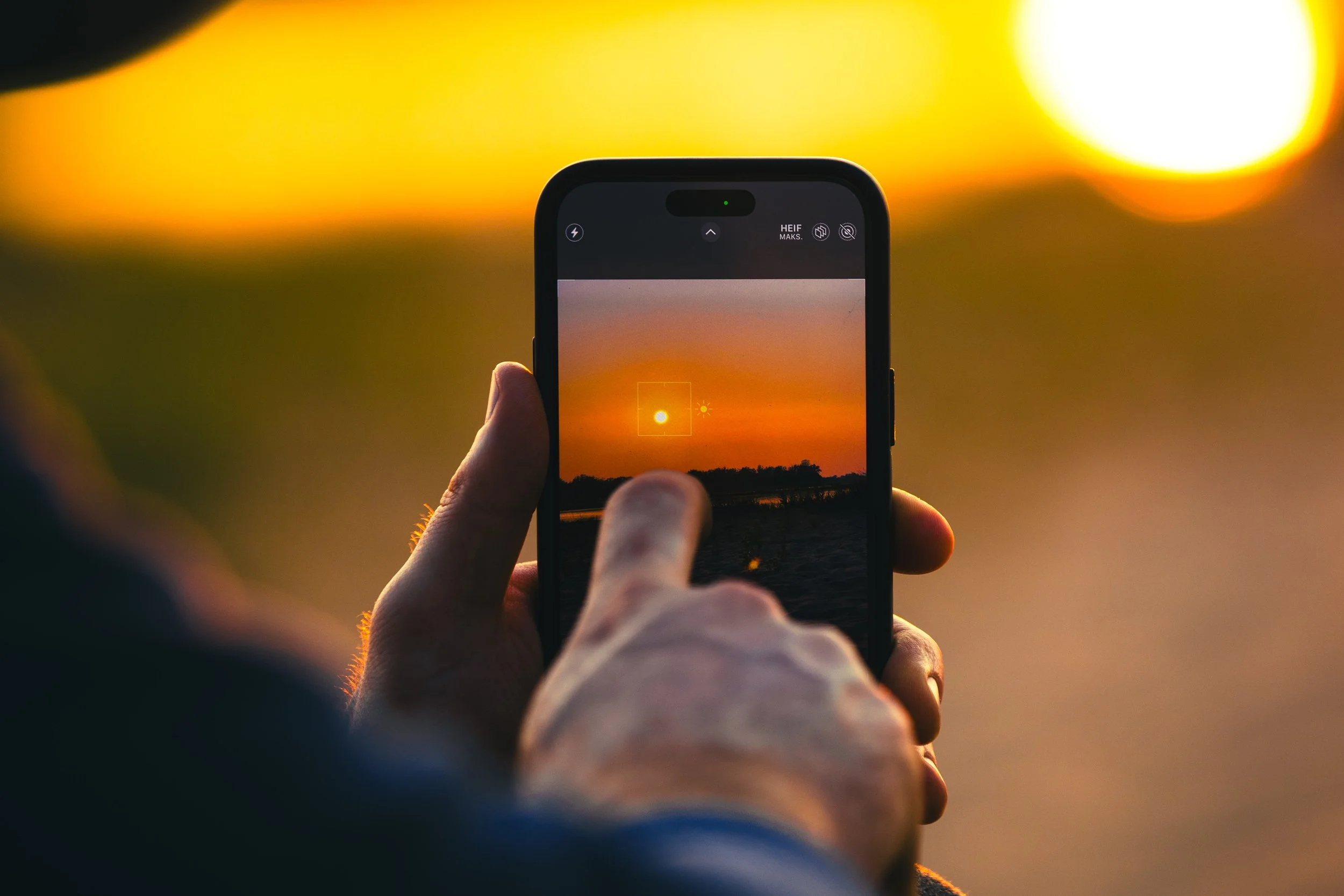 Person taking a photo of a sunset over a lake with a smartphone.