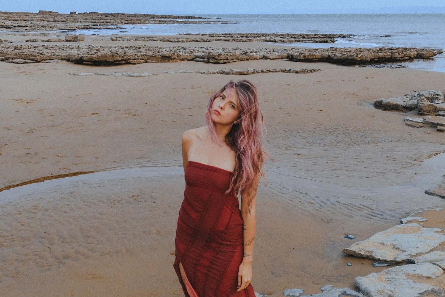A woman with long, wavy pink hair standing on a beach with sand and rocks, wearing a strapless red dress, with water and the ocean in the background.