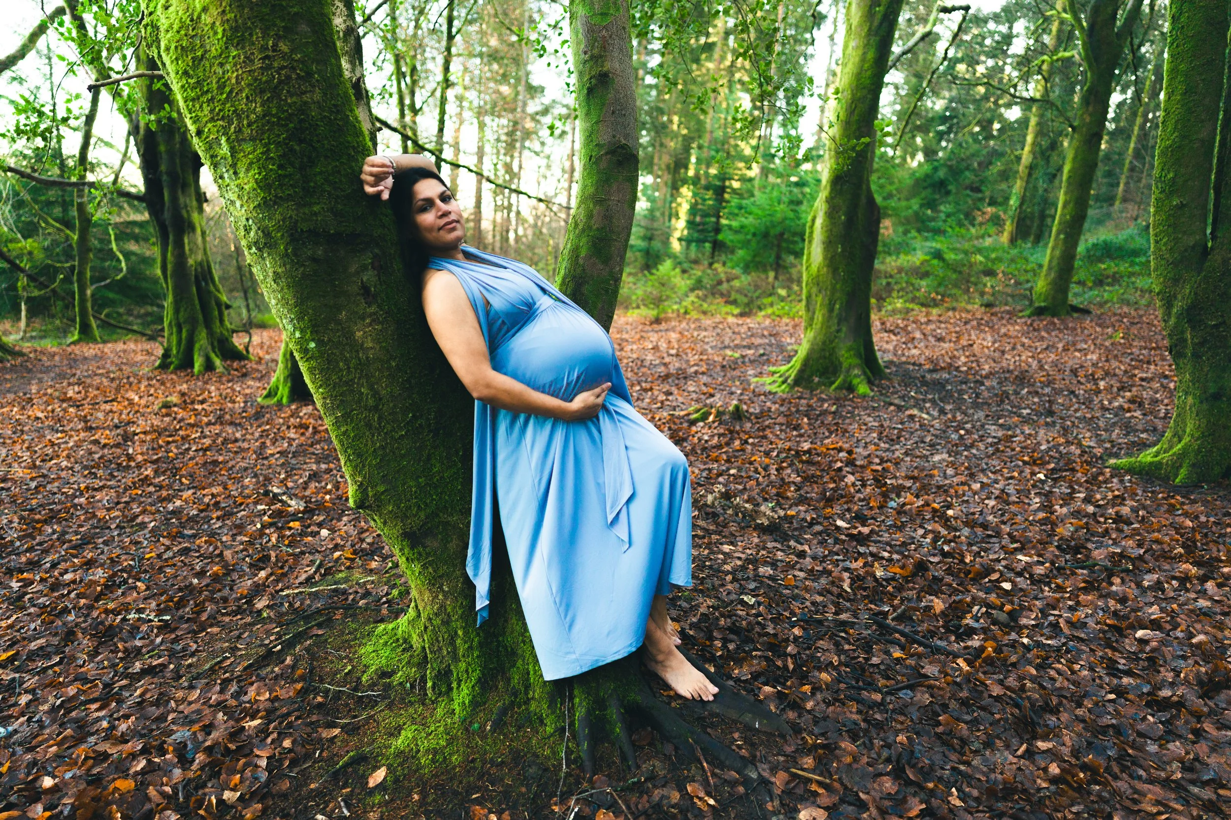 A pregnant woman in a blue dress leaning against a moss-covered tree in a green forest.