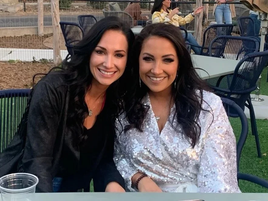 Two women sitting outdoors at a table, smiling at the camera, with empty chairs and a person in the background.