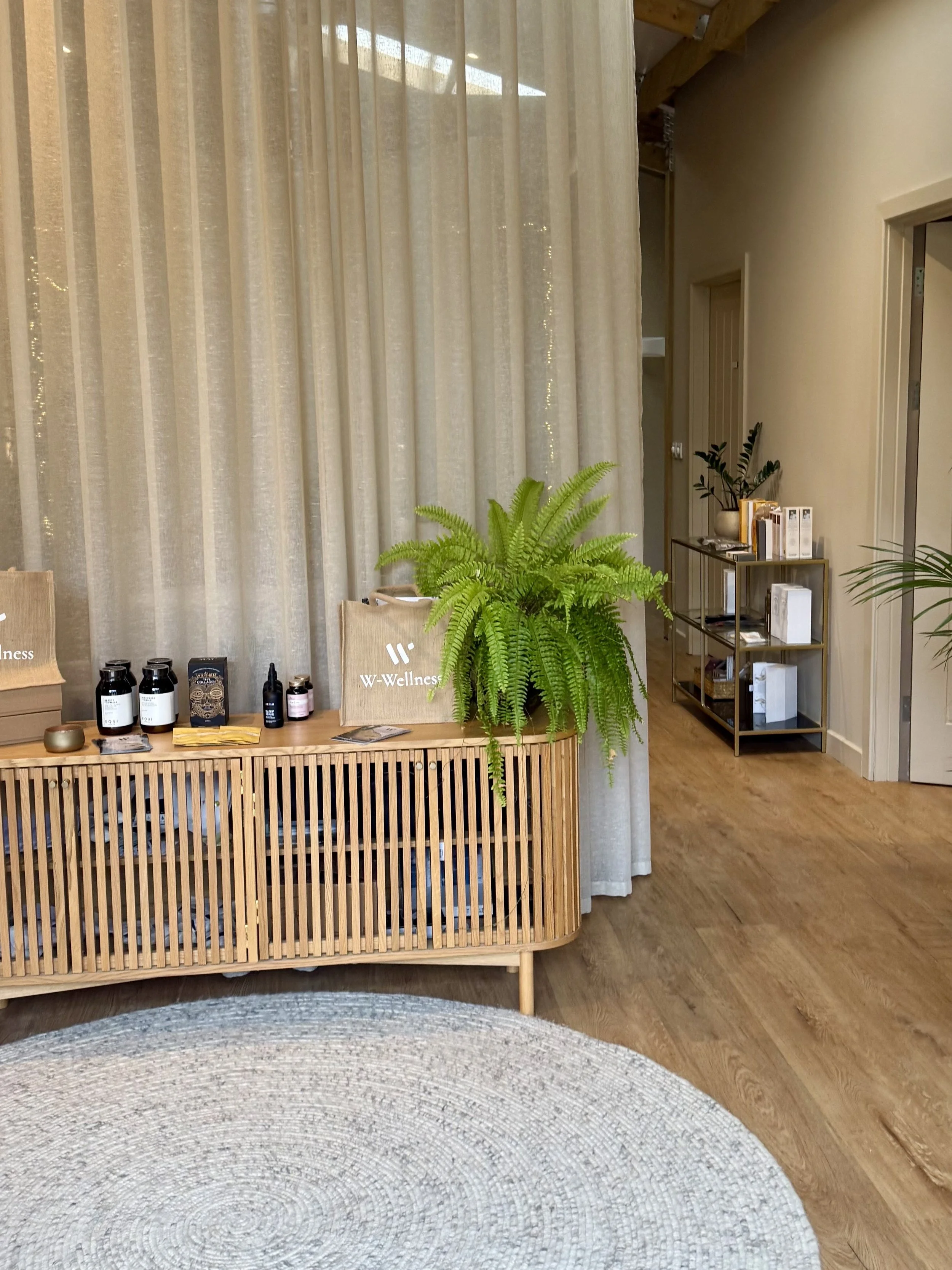 Interior of a wellness center with a wooden reception desk, a large potted fern, and shelves with books and plants. Light-colored curtains hang in the background, and the flooring is wooden with a round woven rug in the foreground.