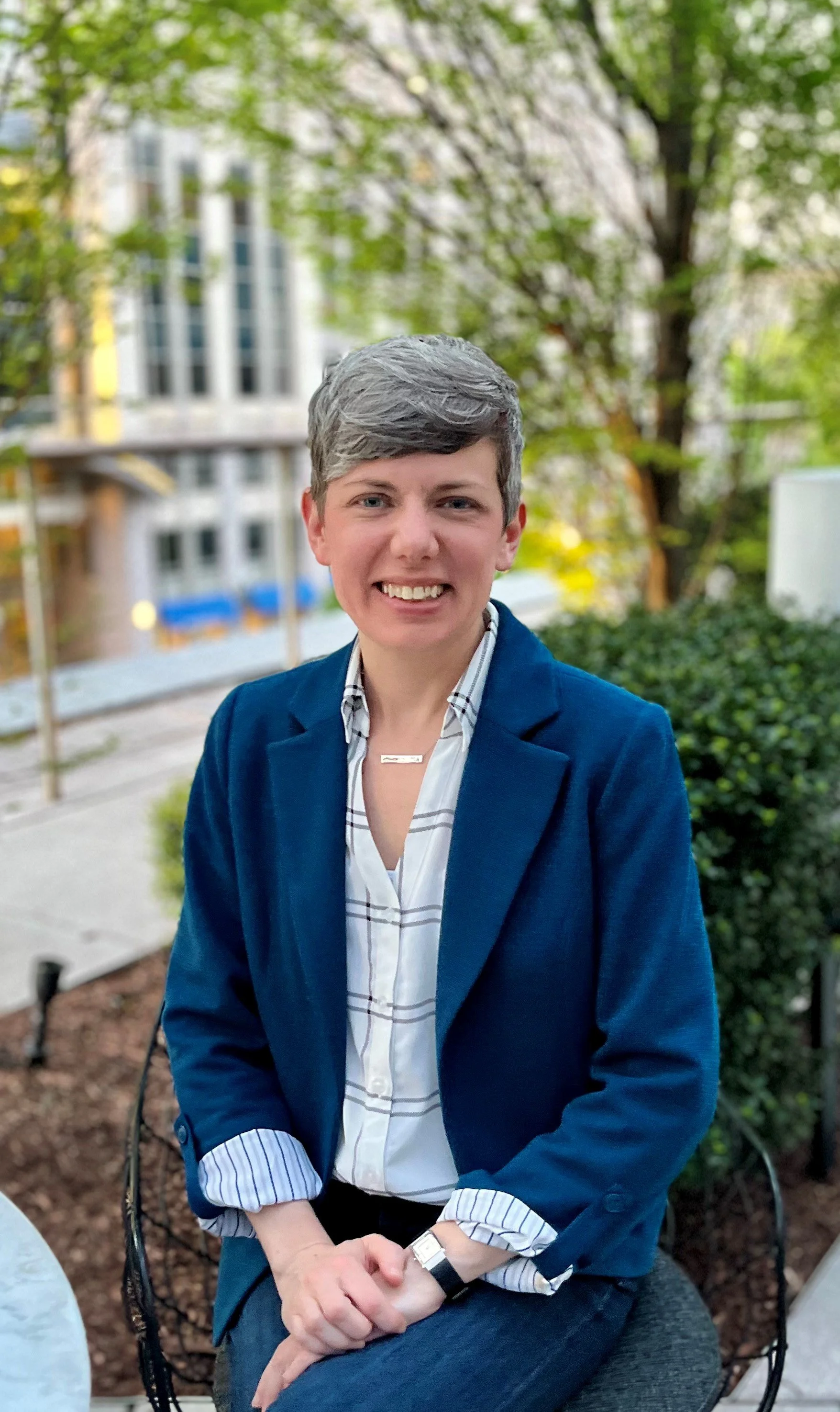 A woman with short gray hair smiling, wearing a dark blue blazer, a light blue striped shirt, and a watch, sitting outdoors at a table with a blurred building and trees in the background.