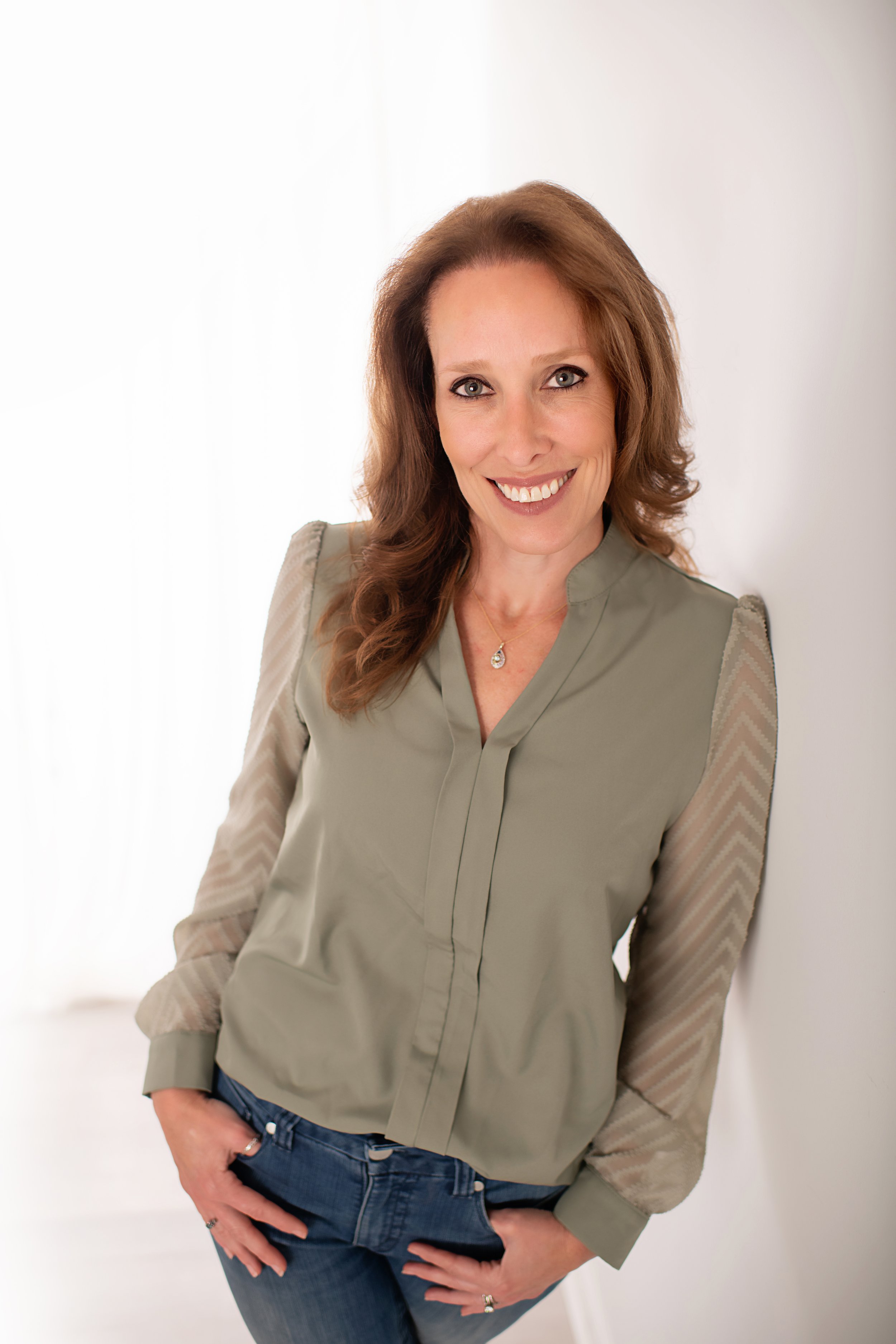 Smiling woman with long brown hair, wearing a light green blouse with sheer sleeves, standing against a white background.