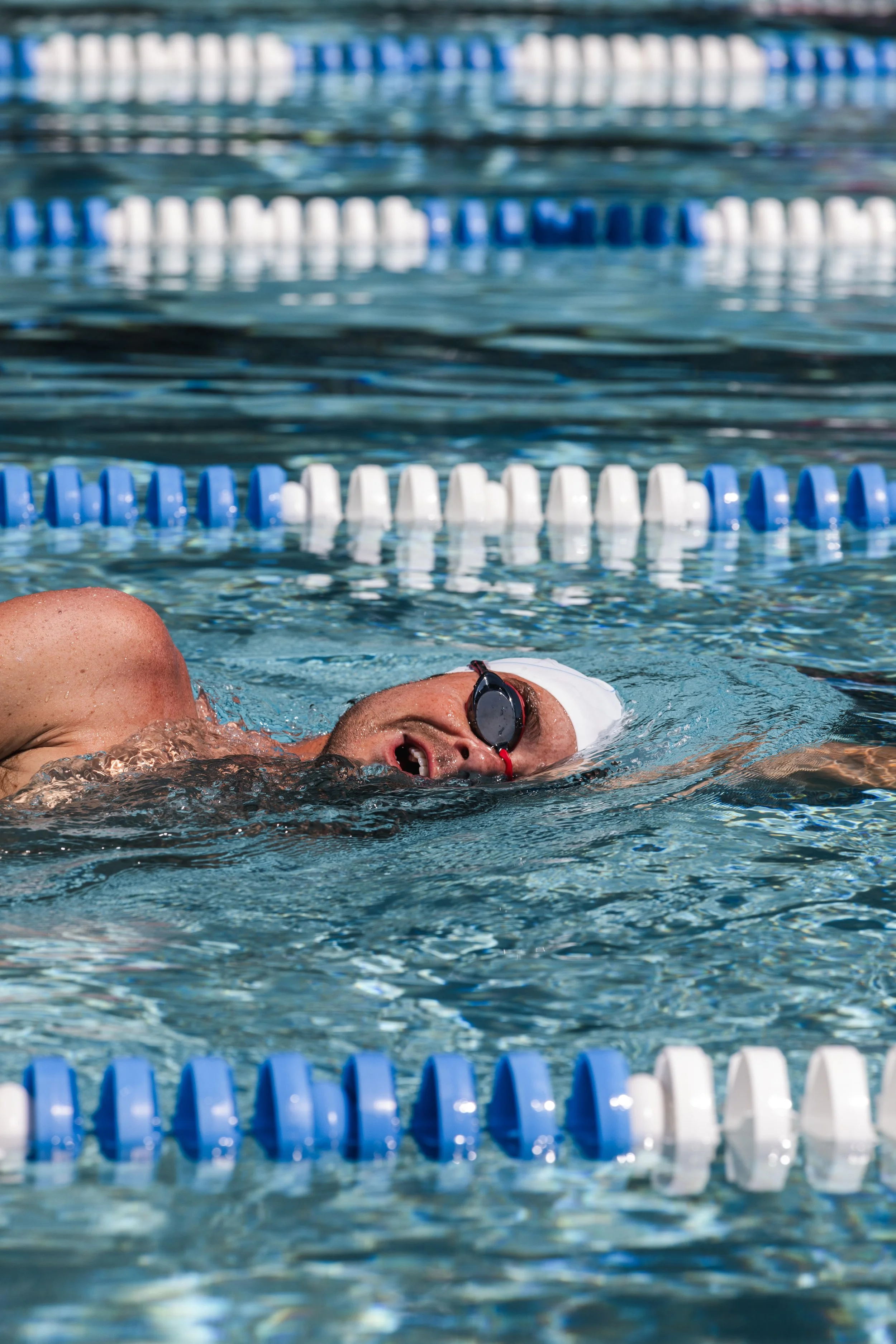 Swimmer in black goggles and white swim cap performing backstroke in an outdoor swimming pool, with blue and white lane dividers.