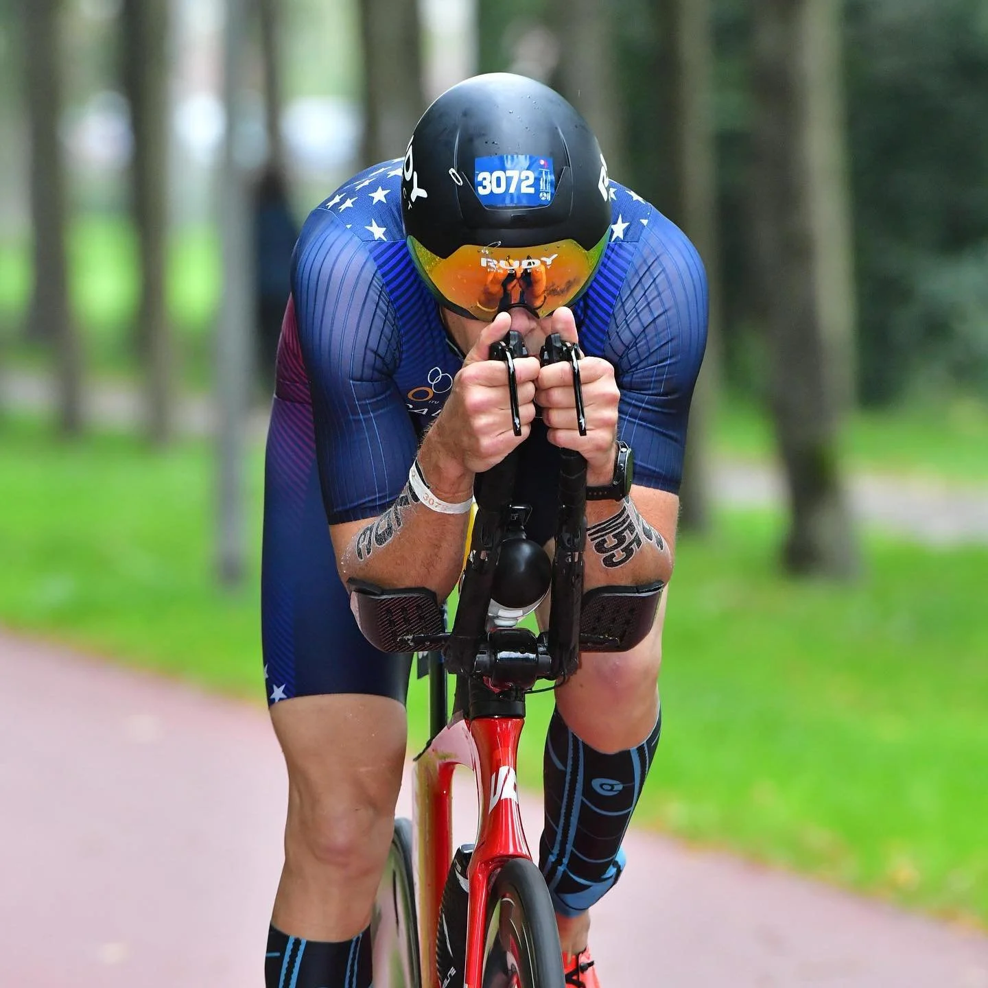 Triathlete in blue and black gear, wearing a helmet with the number 3072, leaning forward on a red bike in a wooded area.