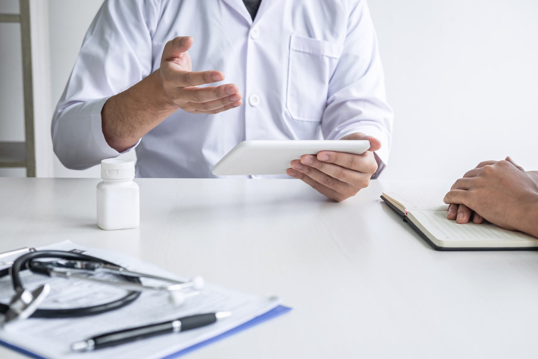 Doctor in white coat showing something on a tablet to a patient, with medical documents and a pill bottle on the table.