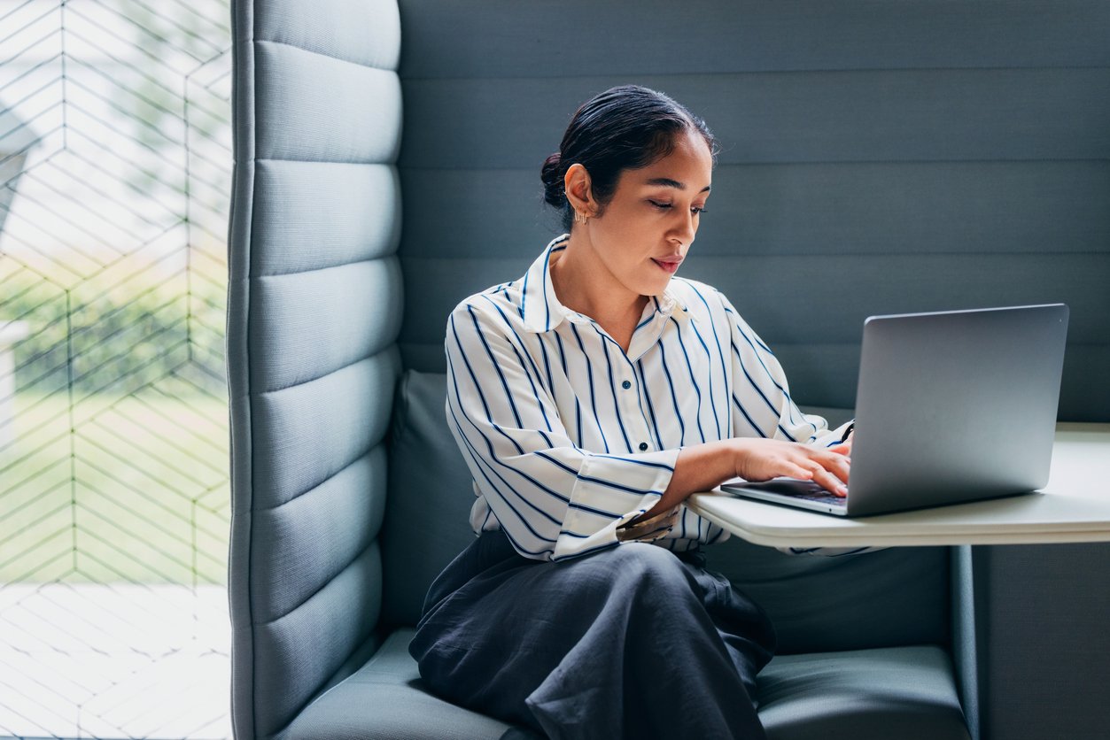A woman sitting in a booth working on a laptop, wearing a white shirt with blue stripes and black pants.
