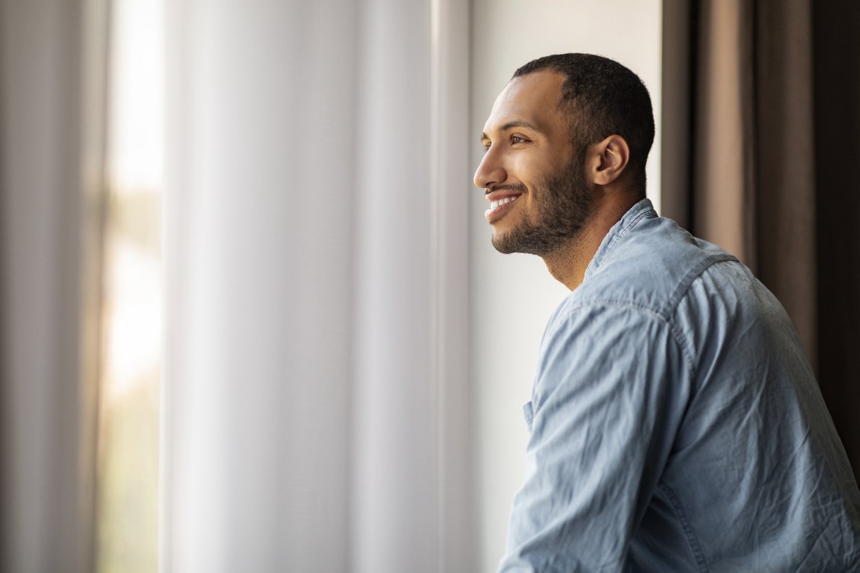 A man with a beard and short dark hair sitting by a window, smiling, wearing a light blue shirt.