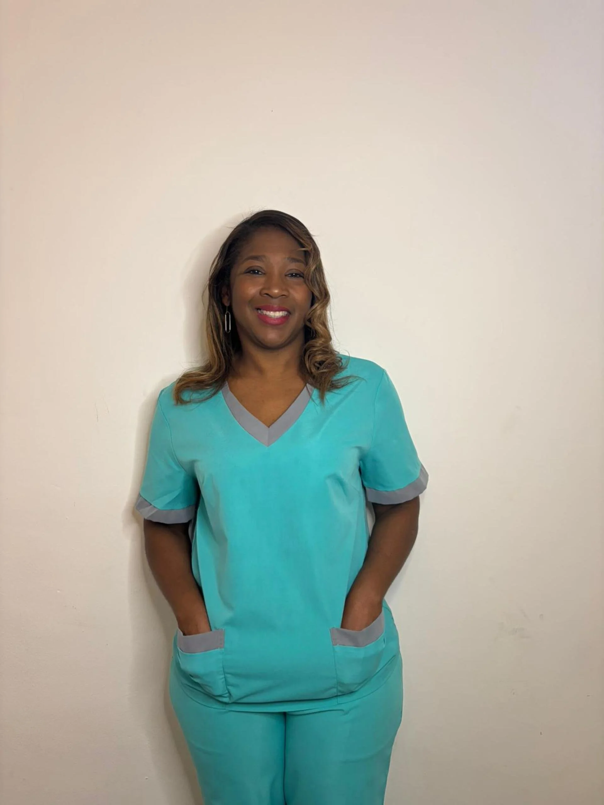 Mujer sonriendo, con uniforme de tipo scrubs en tonos turquoise con detalles grises, de pie junto a una pared blanca.