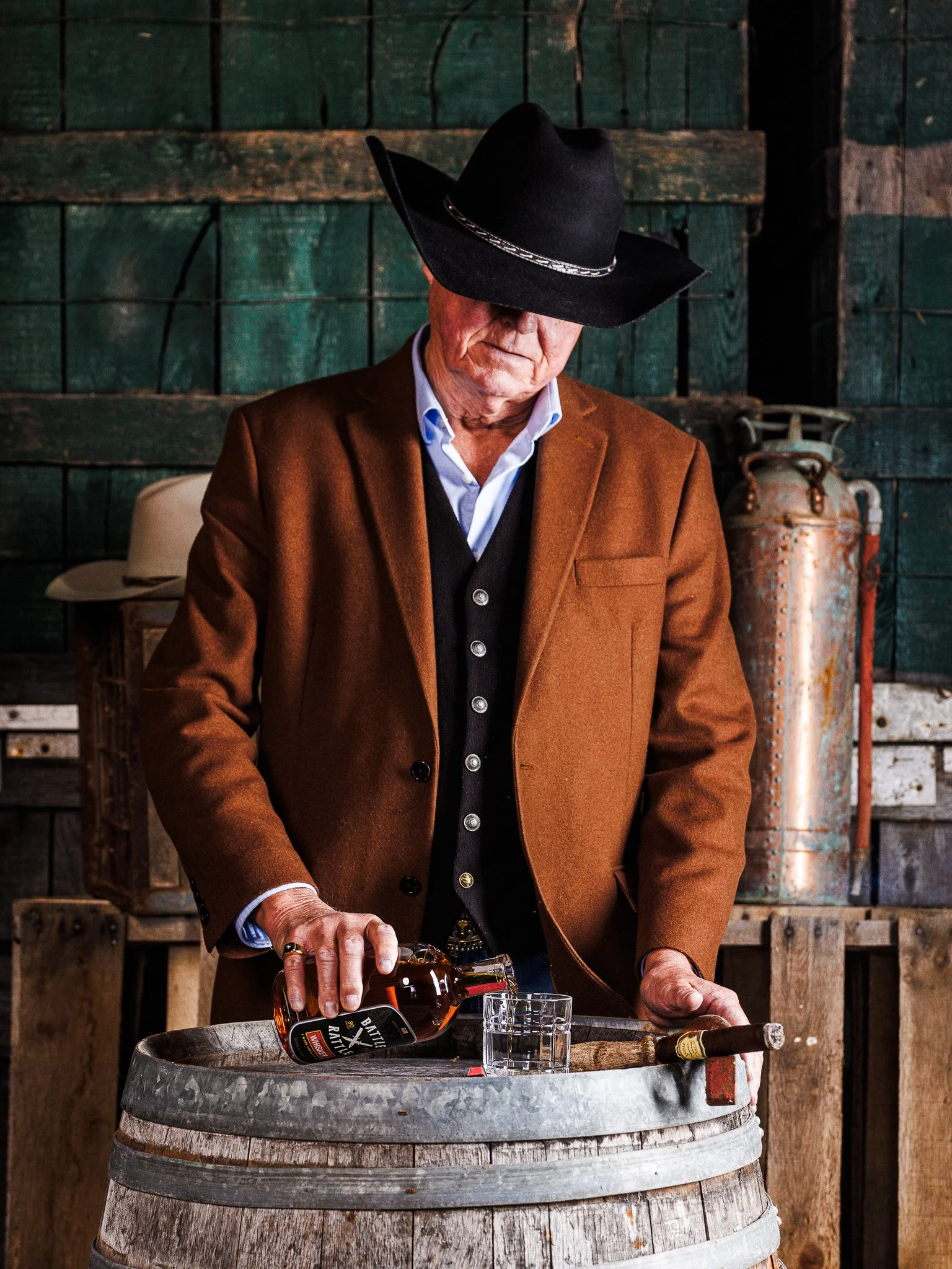 An older man wearing a black cowboy hat, brown blazer, and blue shirt pours whisky into a glass placed on a wooden barrel, with a cigar in hand, in a rustic setting with wooden walls and vintage items.