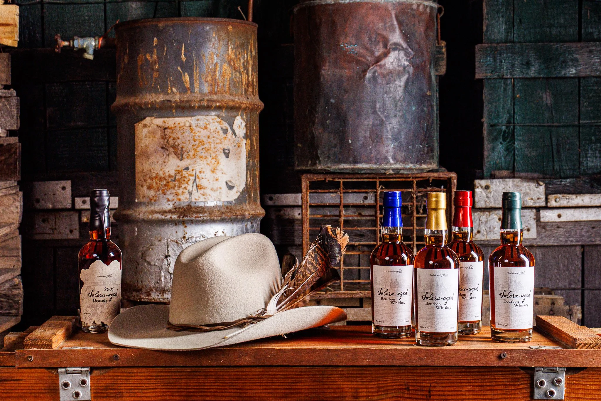 Four bottles of Solera-aged bourbon whiskey, a beige cowboy hat with feathers, a bottle of 2005 Solera-aged brandy, set on a rustic wooden table in a vintage industrial setting with rusty drums and wooden planks.