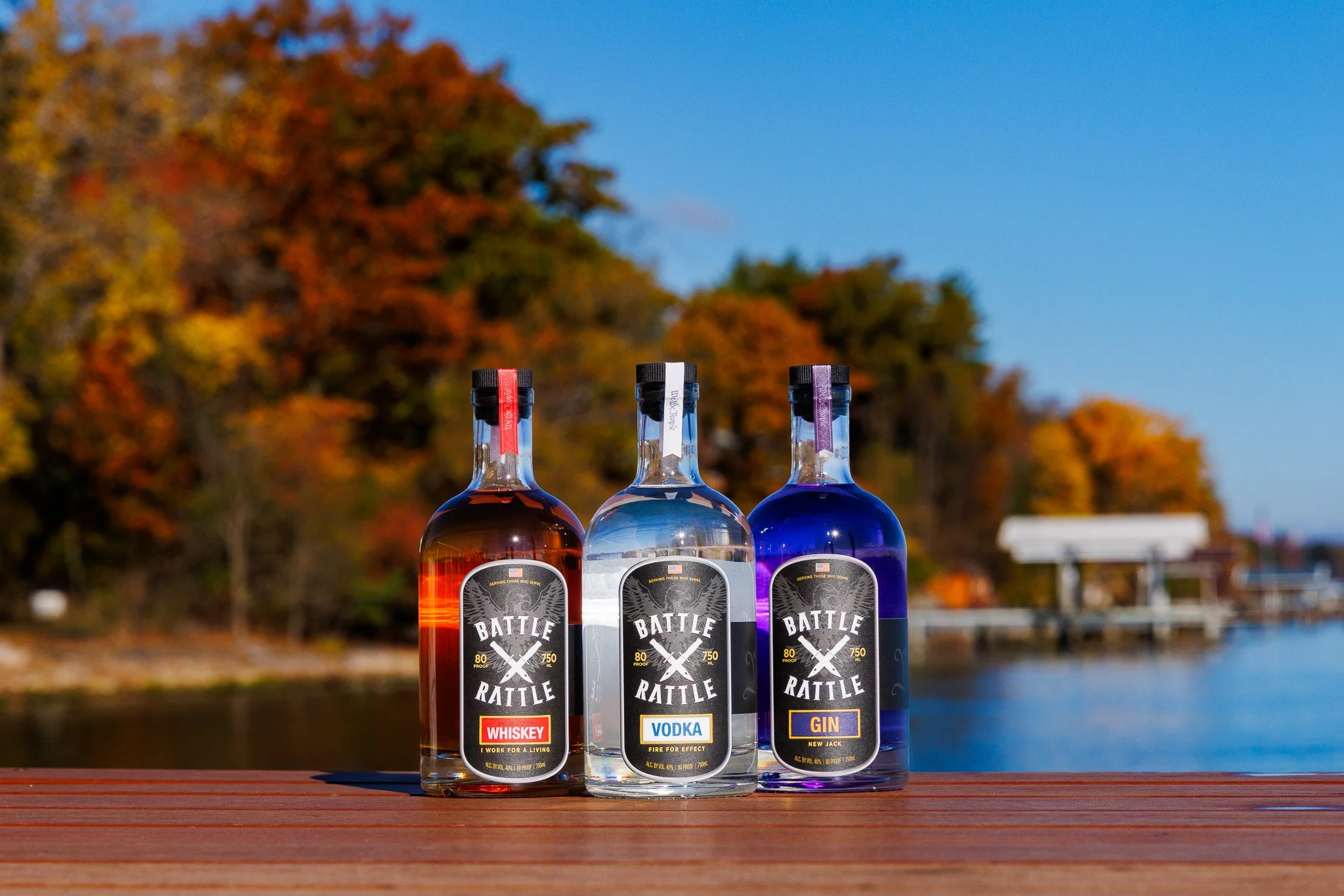Three bottles of flavored spirits, whiskey, vodka, and gin, on a wooden surface outdoors with a river and colorful autumn trees in the background.