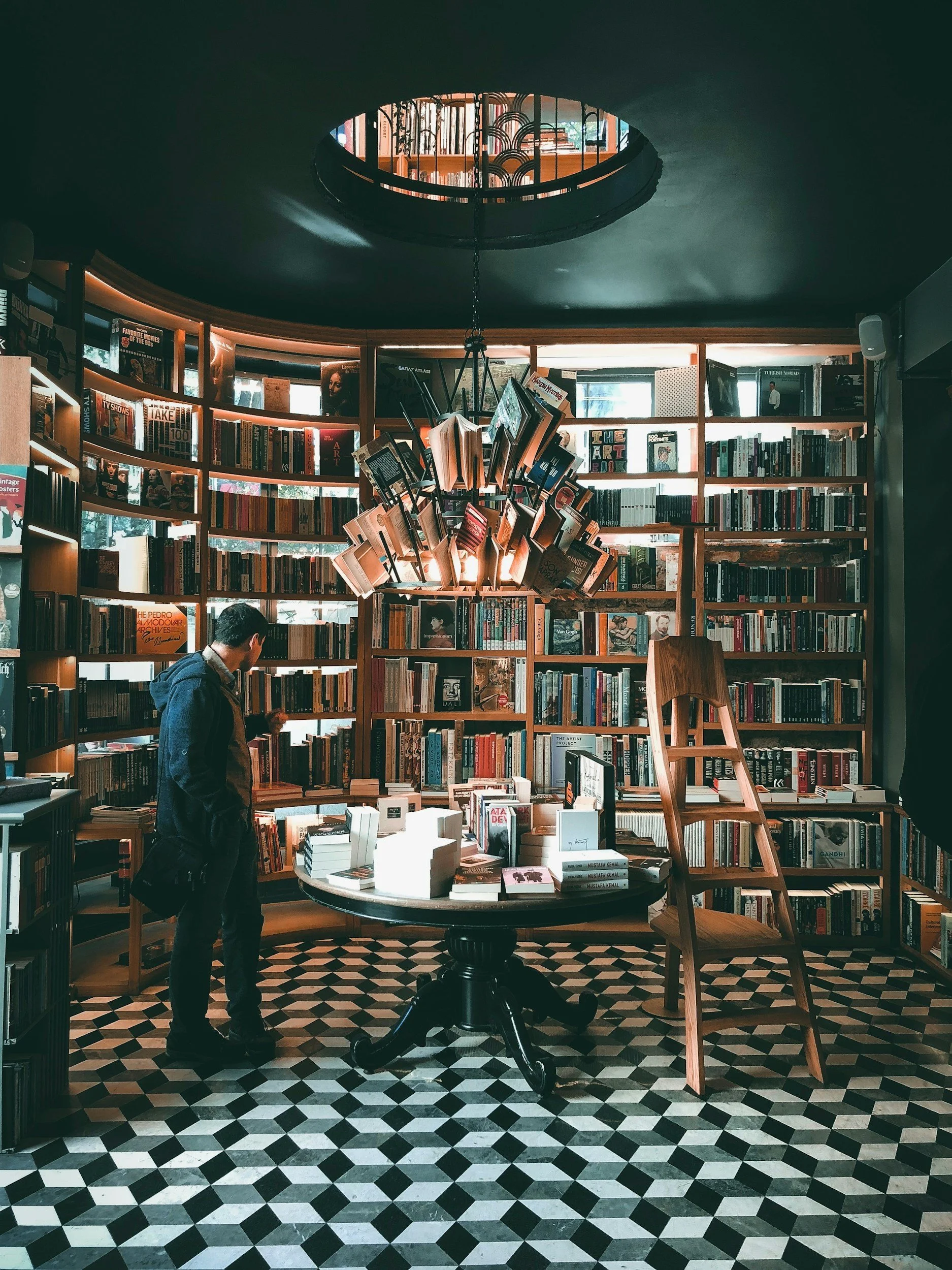 A person browsing books in a bookstore with curved bookshelves, a central round table, a unique chandelier made of open books, and black-and-white checkered floor.