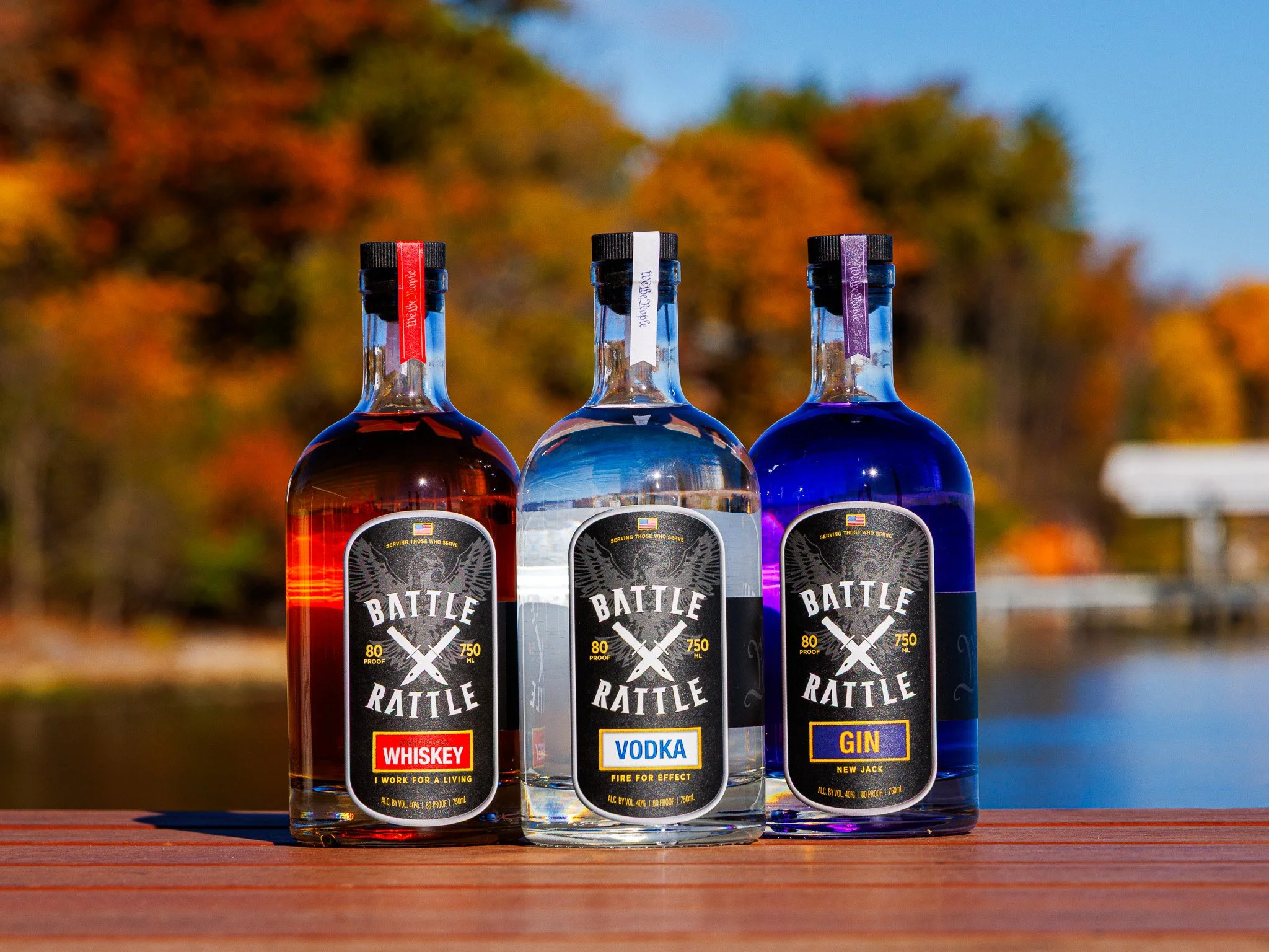 Three bottles of flavored alcohol (whiskey, vodka, gin) on a wooden surface with autumn-colored trees in the background.