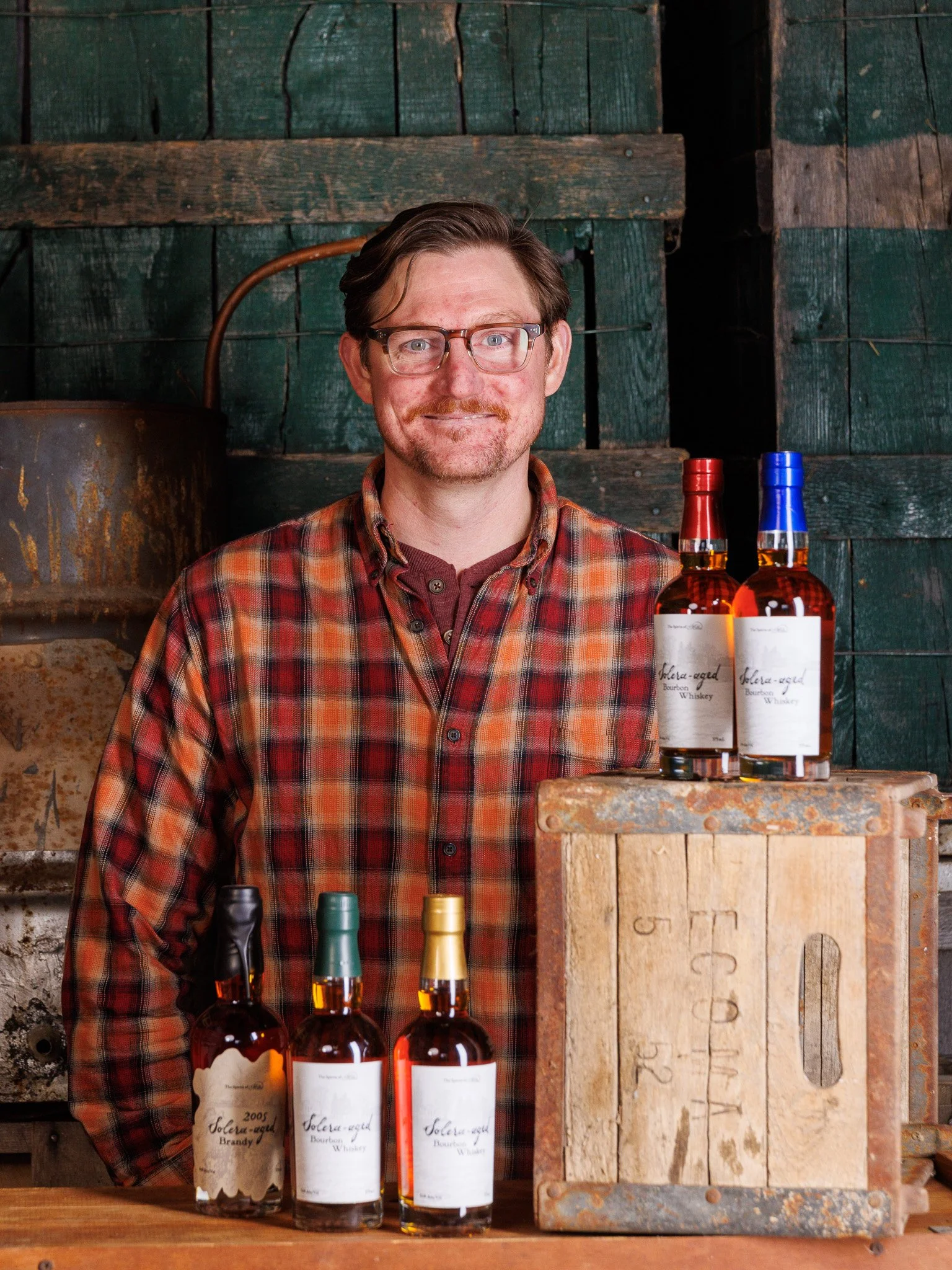 Man with glasses and a plaid shirt standing behind bottles of bourbon whiskey in a rustic wood setting.