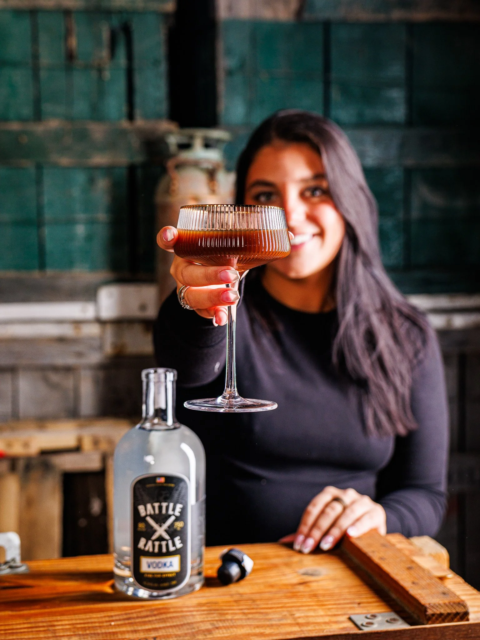 A woman with long dark hair is holding a cocktail glass filled with a dark drink, extending it toward the camera. There is a bottle of vodka on the wooden table in front of her, with a rustic background.
