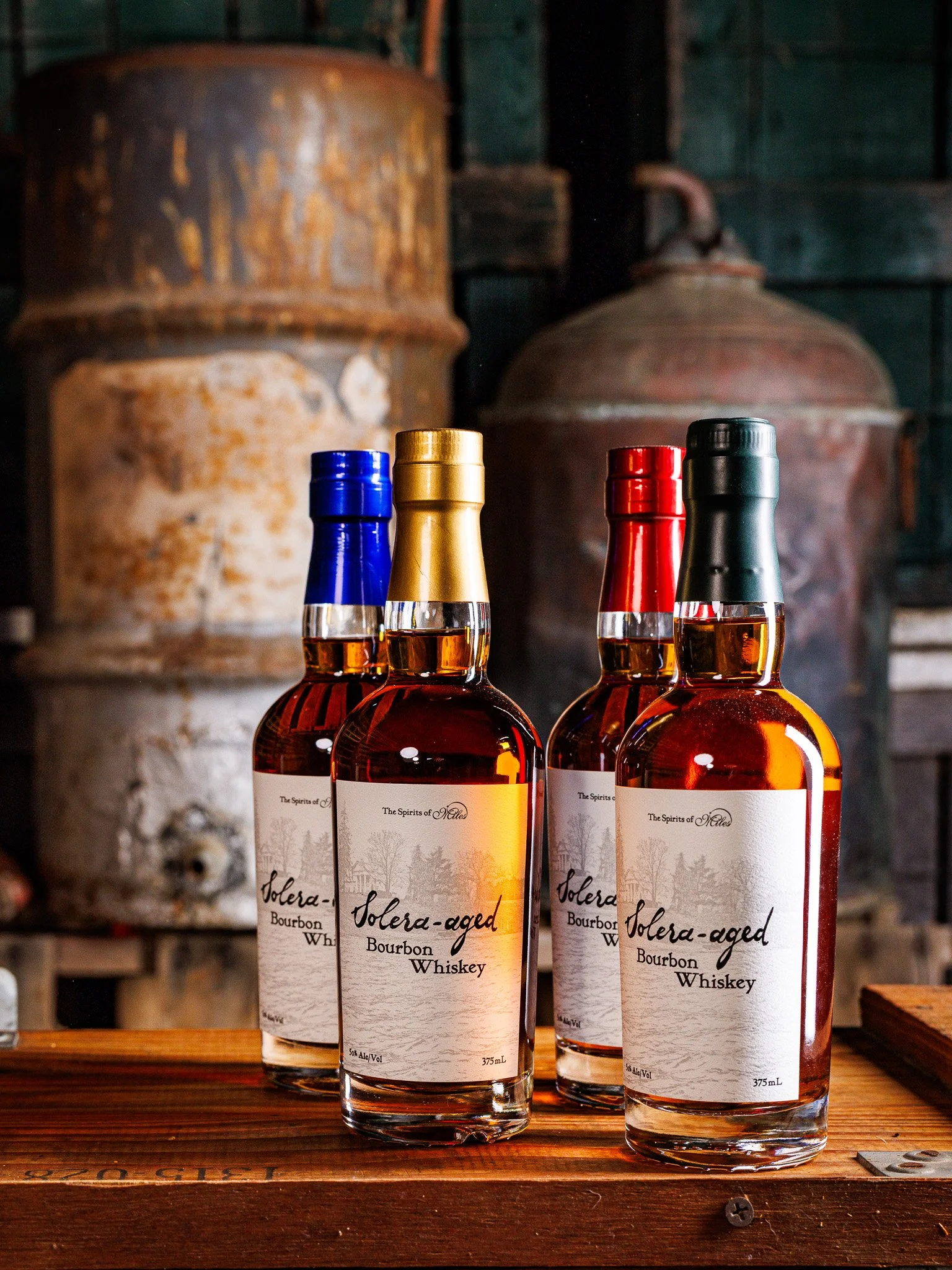 Four bottles of Solera-aged bourbon whiskey with colored caps (blue, gold, red, black) on a wooden surface in front of rustic metal tanks.