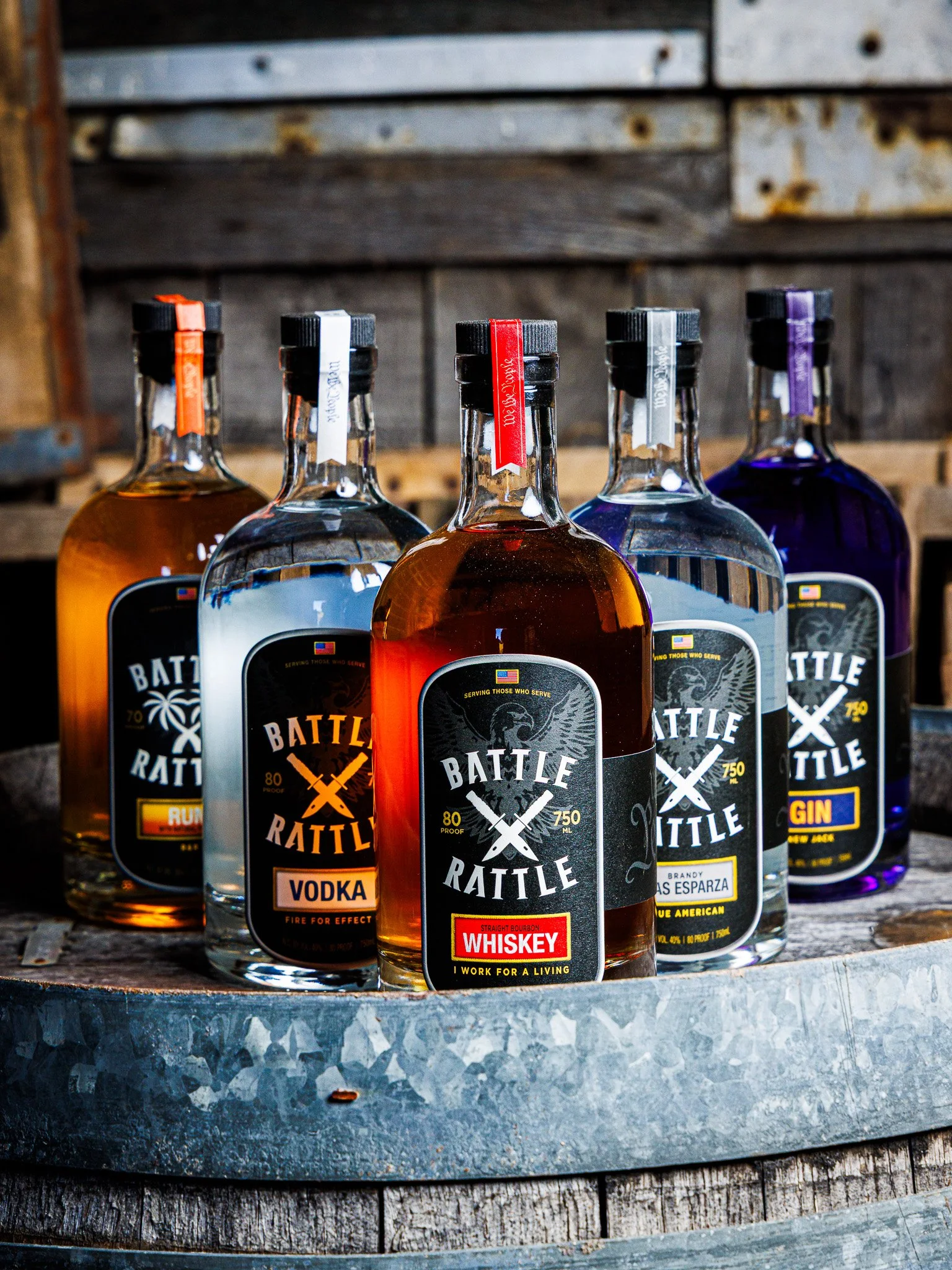 Five bottles of beverage labeled 'Battle Rattle' with different flavors, including whiskey, vodka, rum, gin, and tequila, arranged on a rustic metal surface with a wooden backdrop.