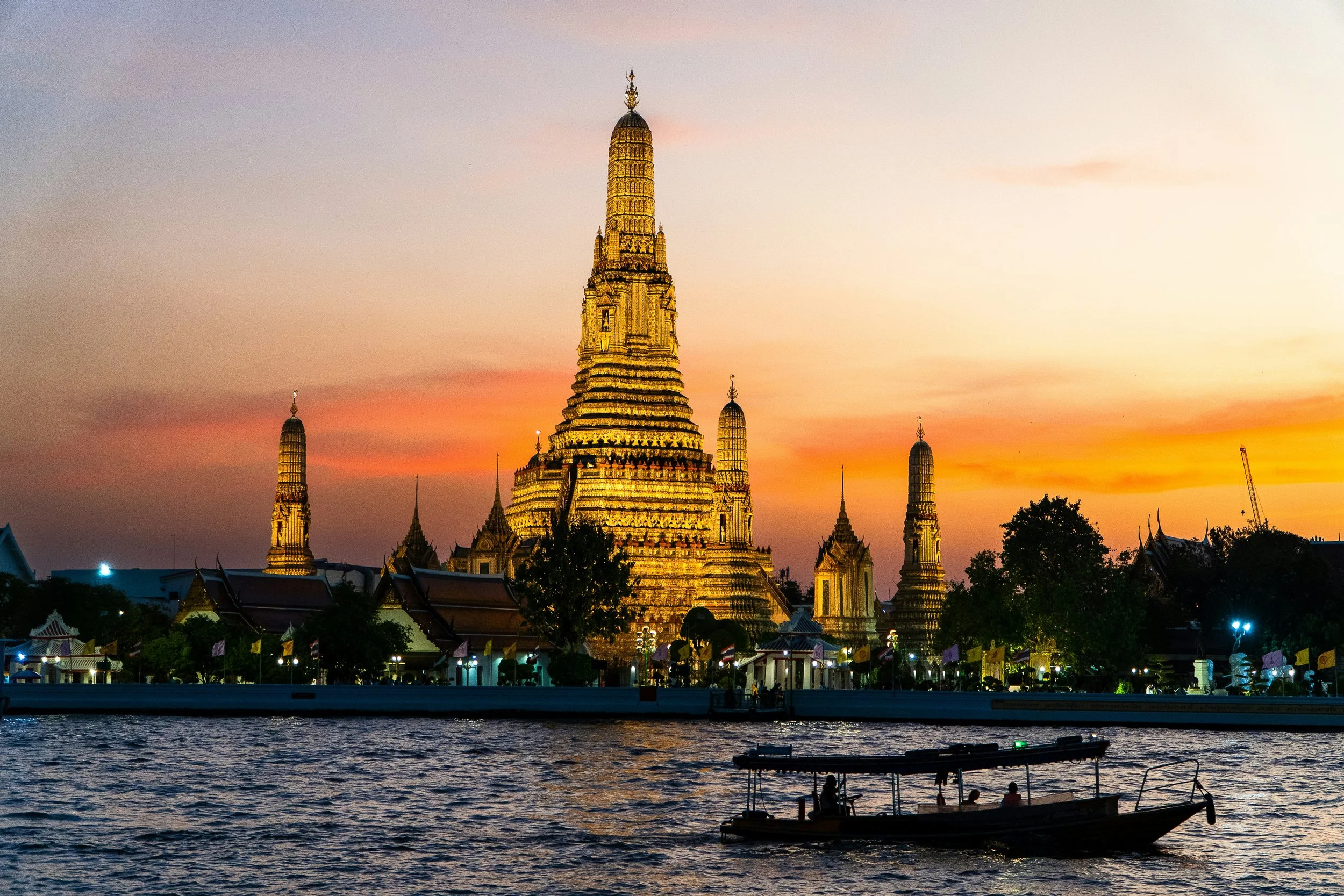 Golden illuminated Wat Arun temple on the Chao Phraya River during sunset in Bangkok, Thailand with boats on the water and a colorful sky.