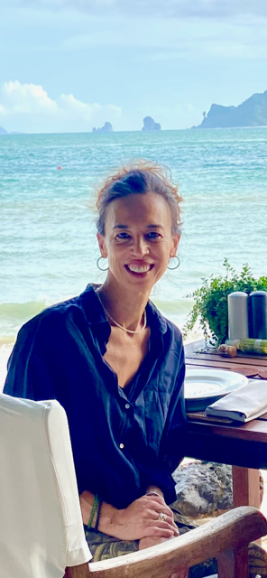 A woman sitting at a table on a beach with a view of the ocean and distant islands, smiling at the camera.
