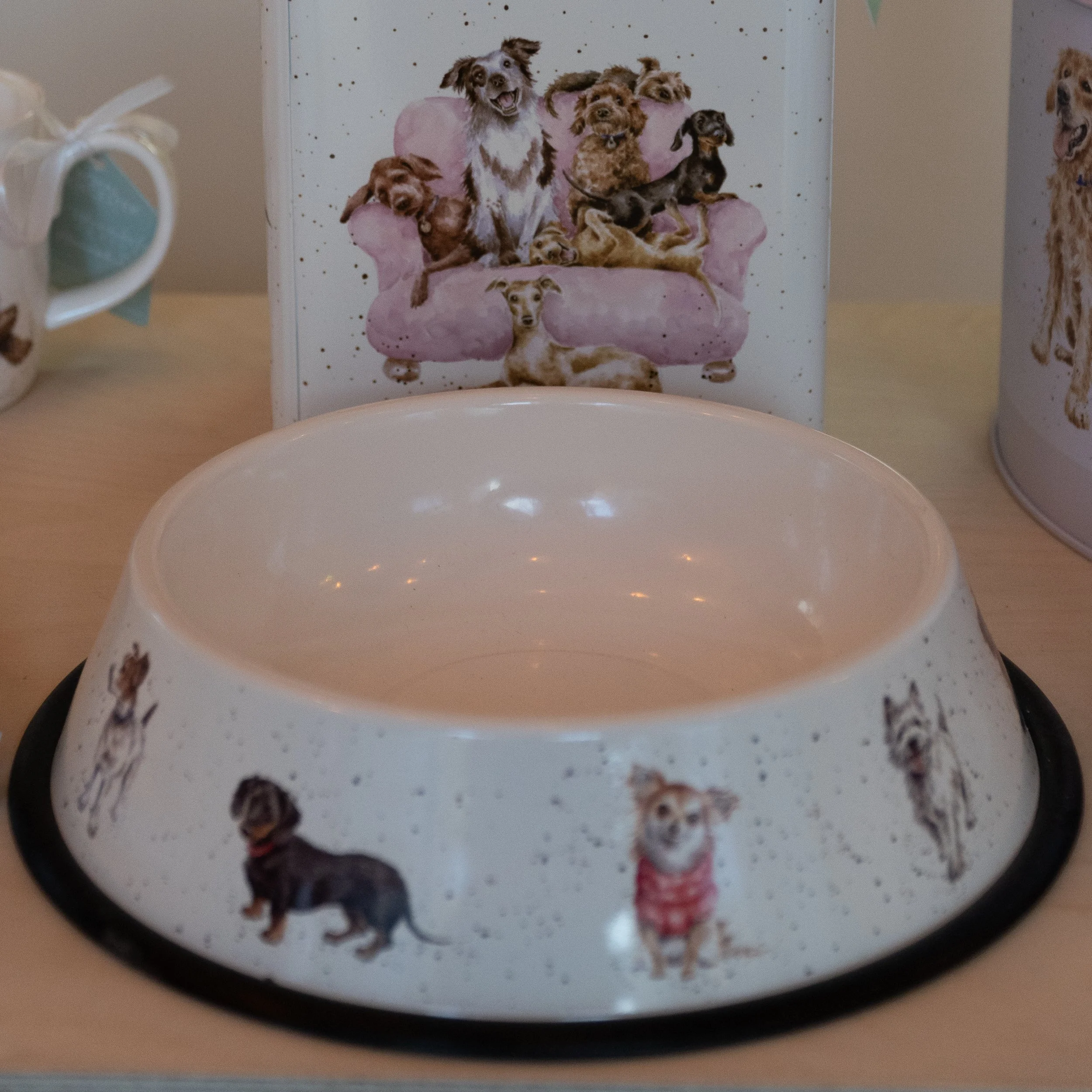 A dog bowl decorated with images of various dog breeds around its border. In the background, there is a box featuring a collage of dogs in different poses, sitting or lying on a pink sofa.