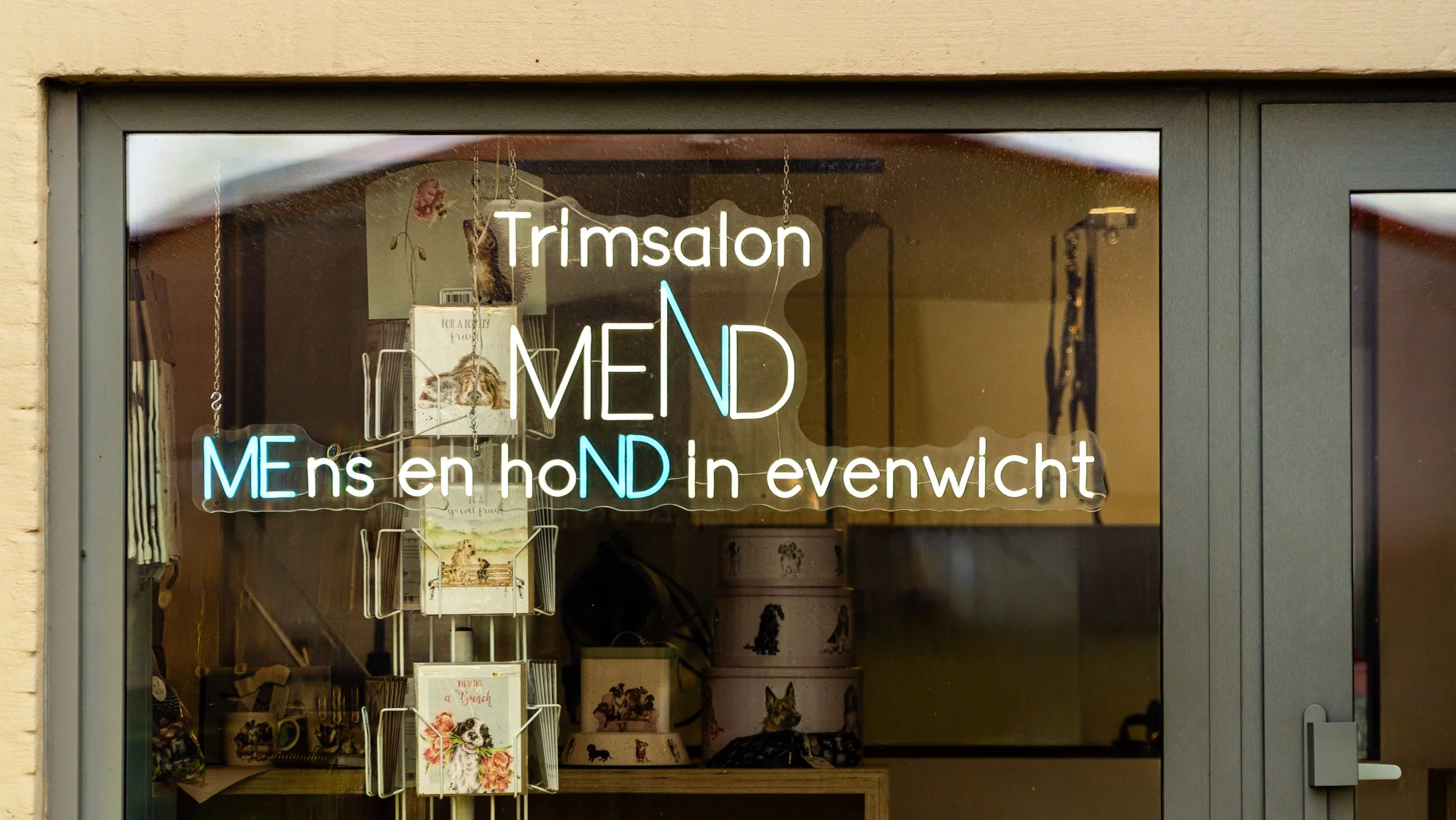 Neon sign for a pet grooming salon with a reflection of the street, displaying the words 'Trimsalon', 'MEND', and 'Mens en hond in evenwicht', with illustrations of cats and dogs in the background.