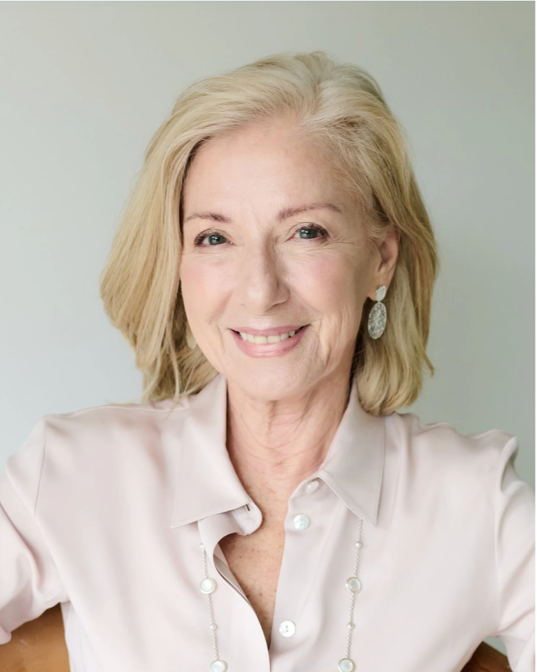 A smiling middle-aged woman with blonde hair, wearing a light-colored blouse with pearl earrings and a matching necklace, posing against a plain light-colored background.