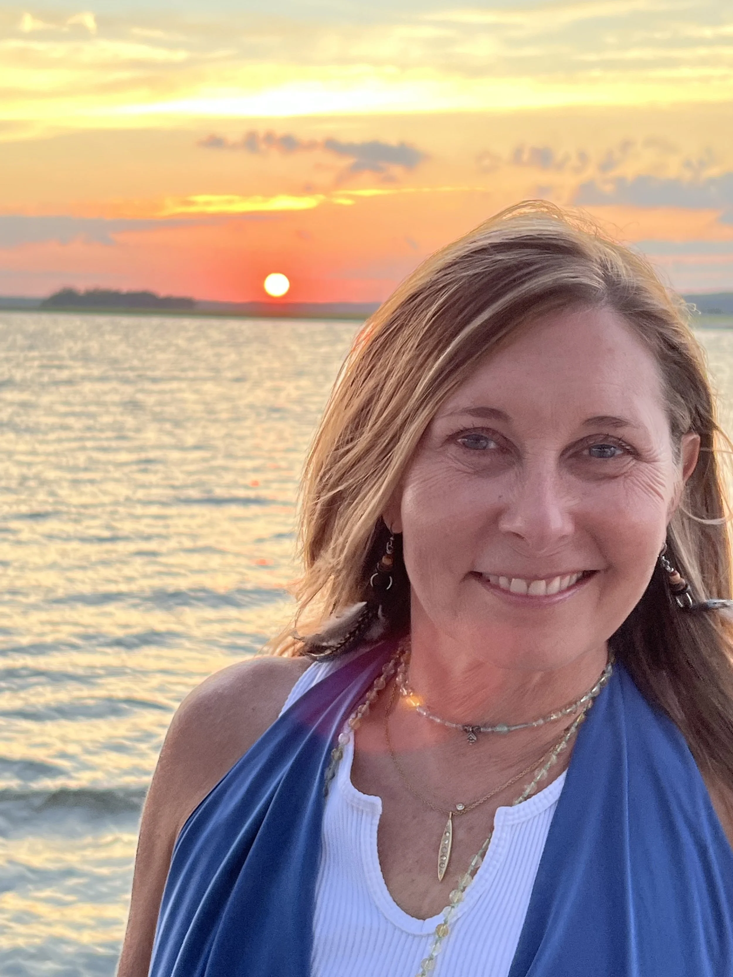 A woman smiling on the beach during sunset with water and a colorful sky in the background.