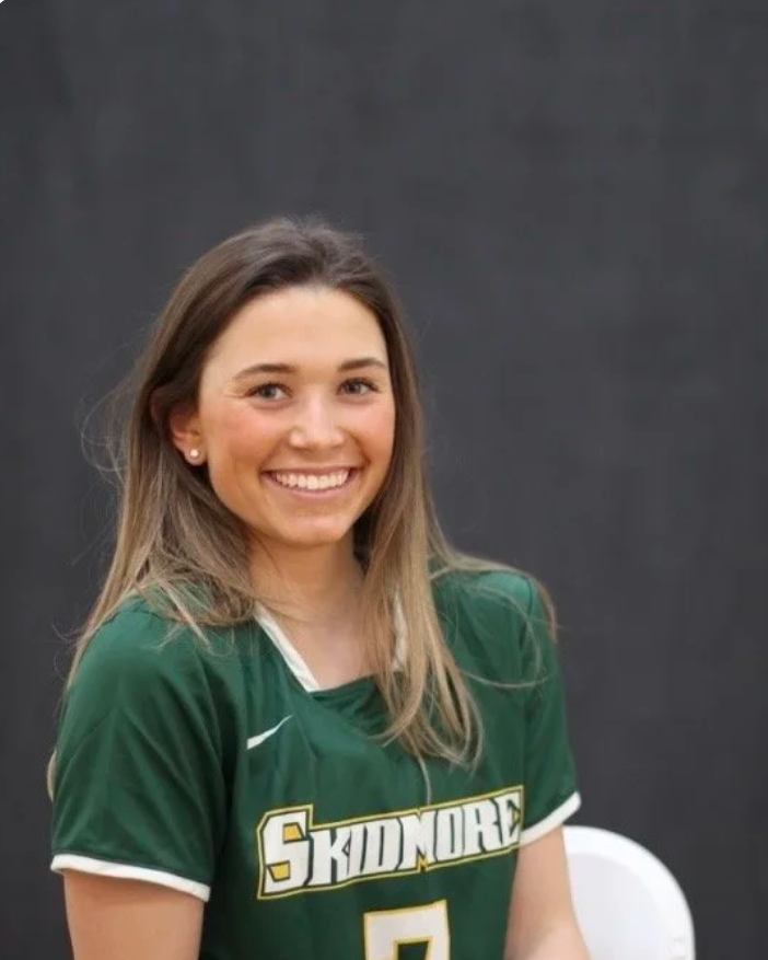 A young woman with long brown hair, wearing a green sports jersey with the word 'SKIDMORE' on it, smiling, sitting against a dark background.