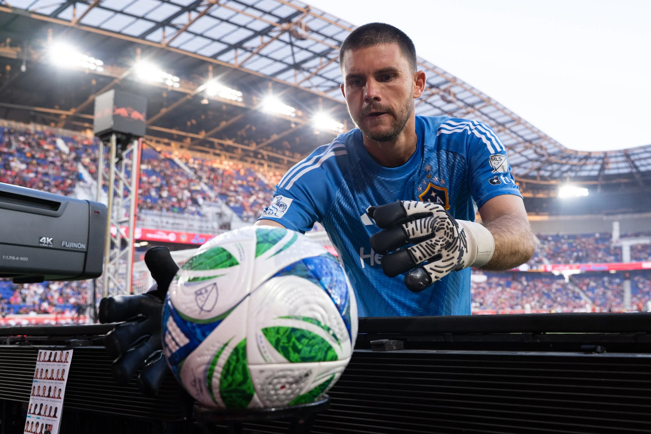 A soccer goalkeeper in a blue uniform preparing to replace a soccer ball. He is wearing gloves and is in a stadium with a crowd in the background.
